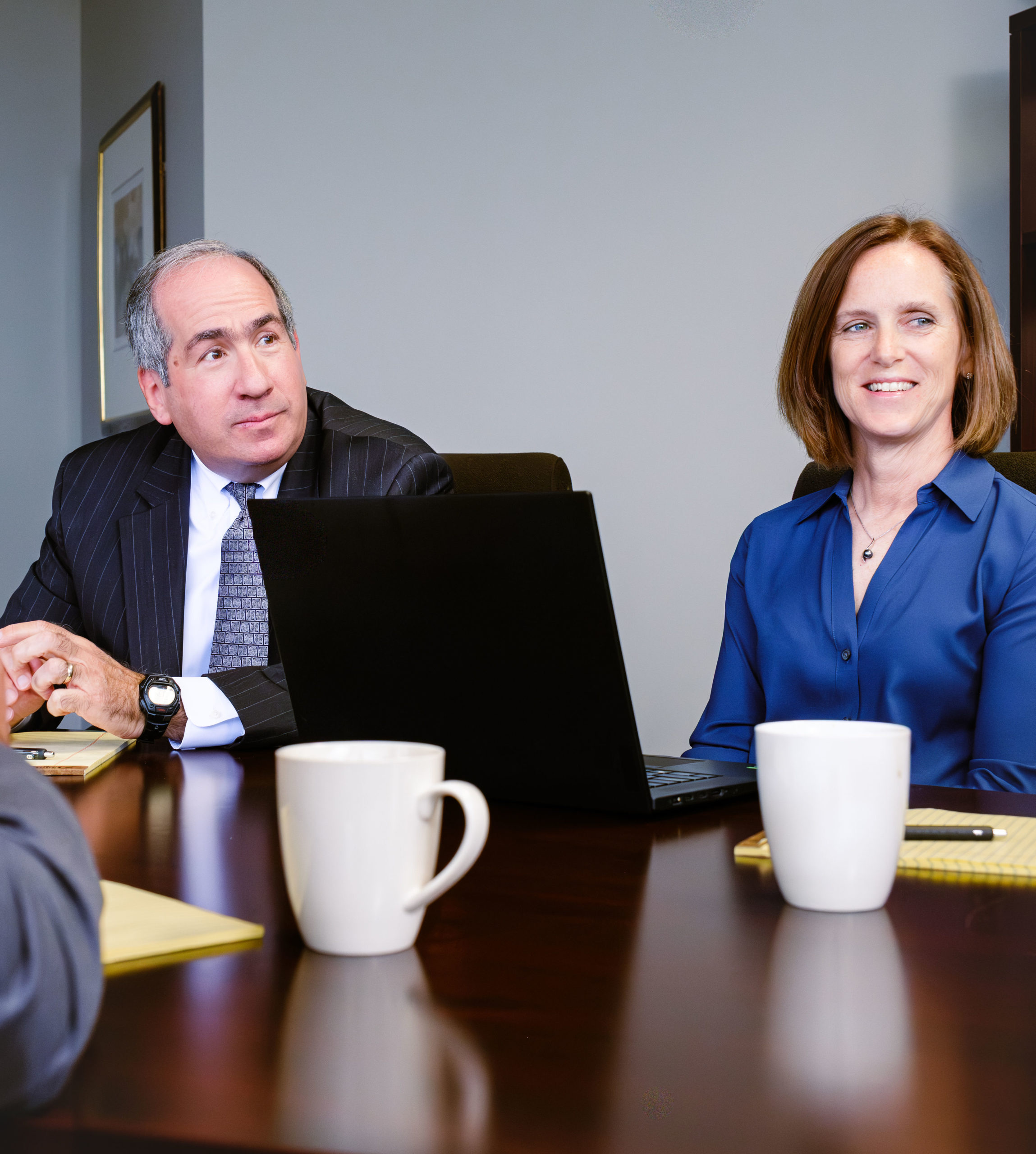 Man in pinstripe suit and woman in blue blouse sitting at conference table with laptop and white coffee mugs during a busi...