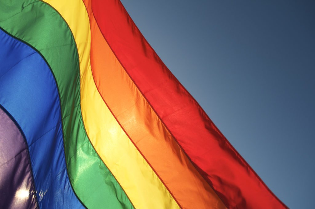 Rainbow flag glows in bright sun waving in blue sky. Some motion blur on top edge of waving flag.