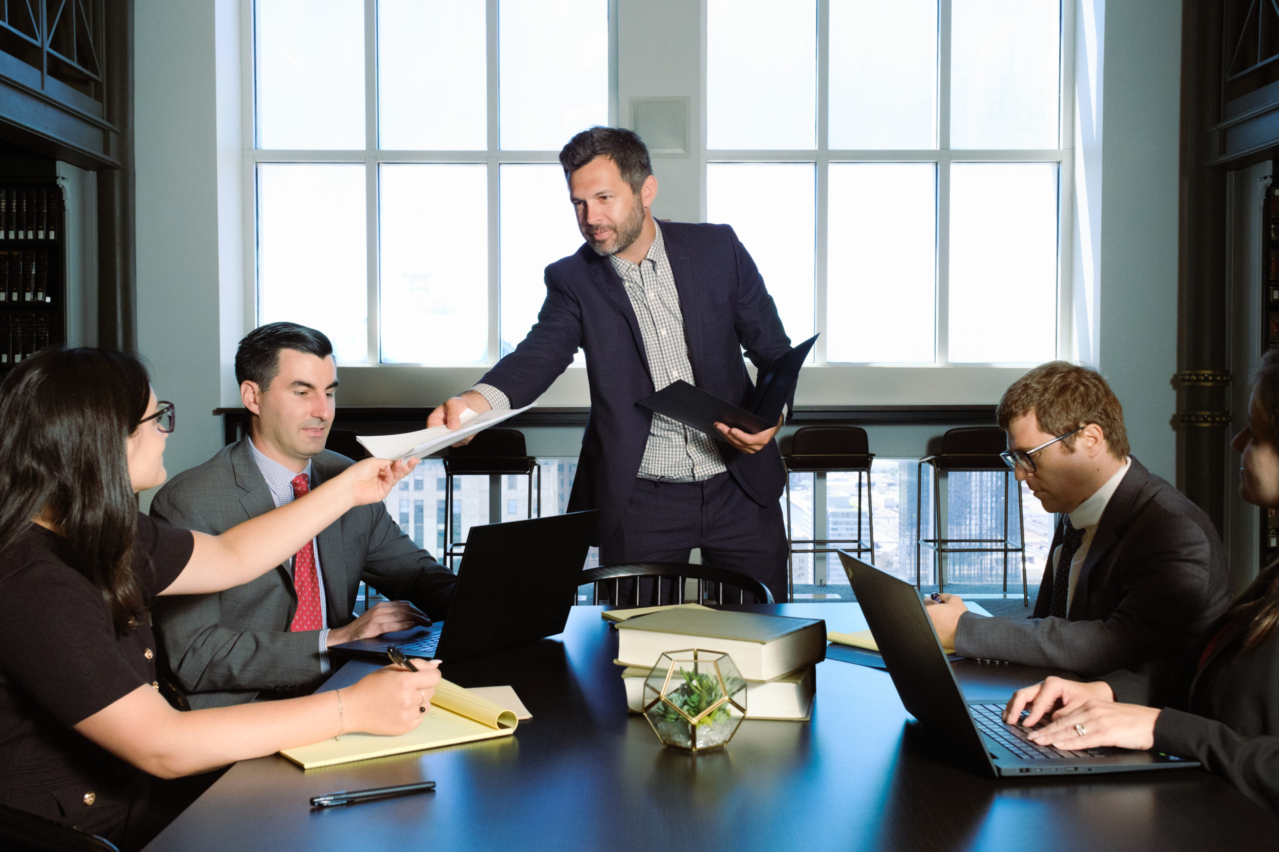 Man in navy suit standing at conference table handing documents to a colleague during a business meeting with four profess...