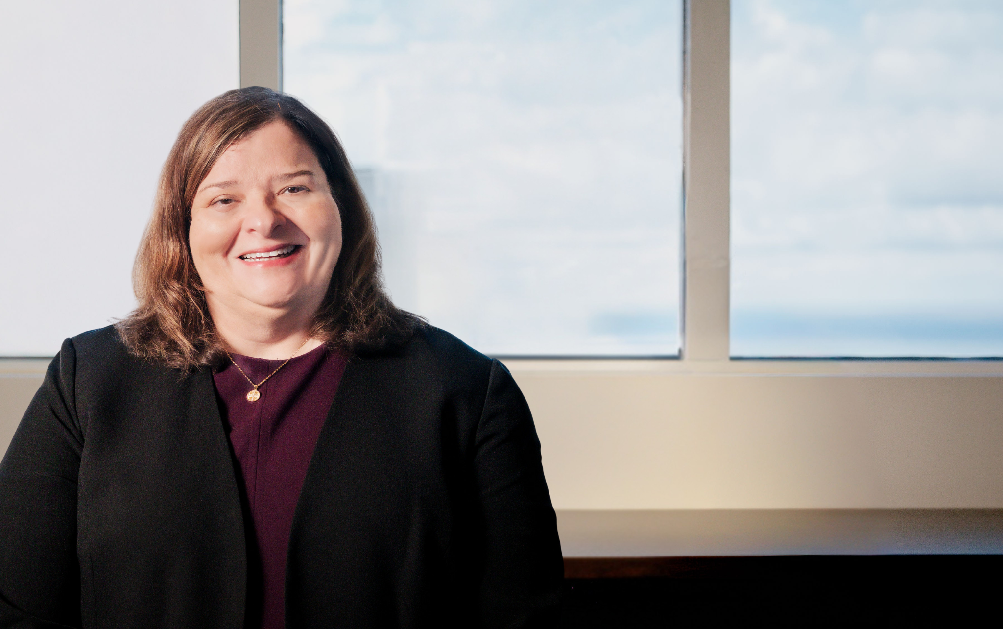 Smiling woman with shoulder-length brown hair wearing a black blazer and burgundy top with a gold necklace, standing near ...