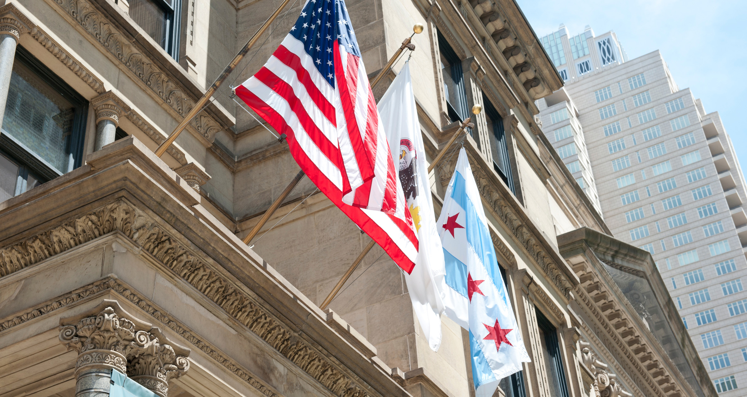 American flag, Illinois state flag, and Chicago city flag displayed on a ornate stone government building with skyscrapers...