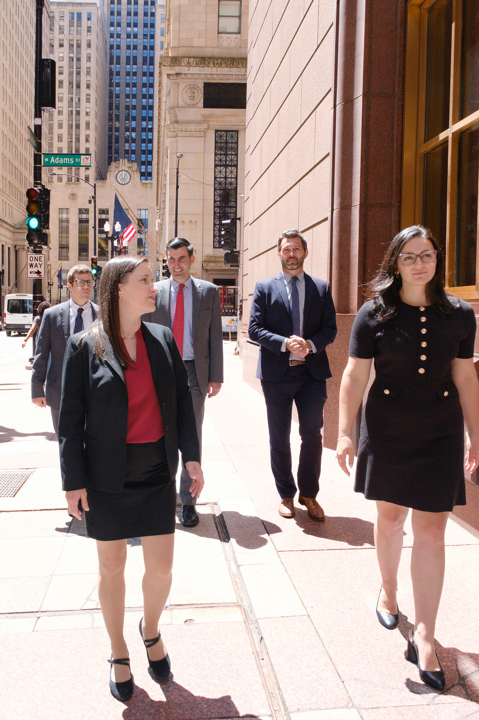 Five business professionals in suits walking on a Chicago downtown sidewalk near W Adams St on a sunny day.