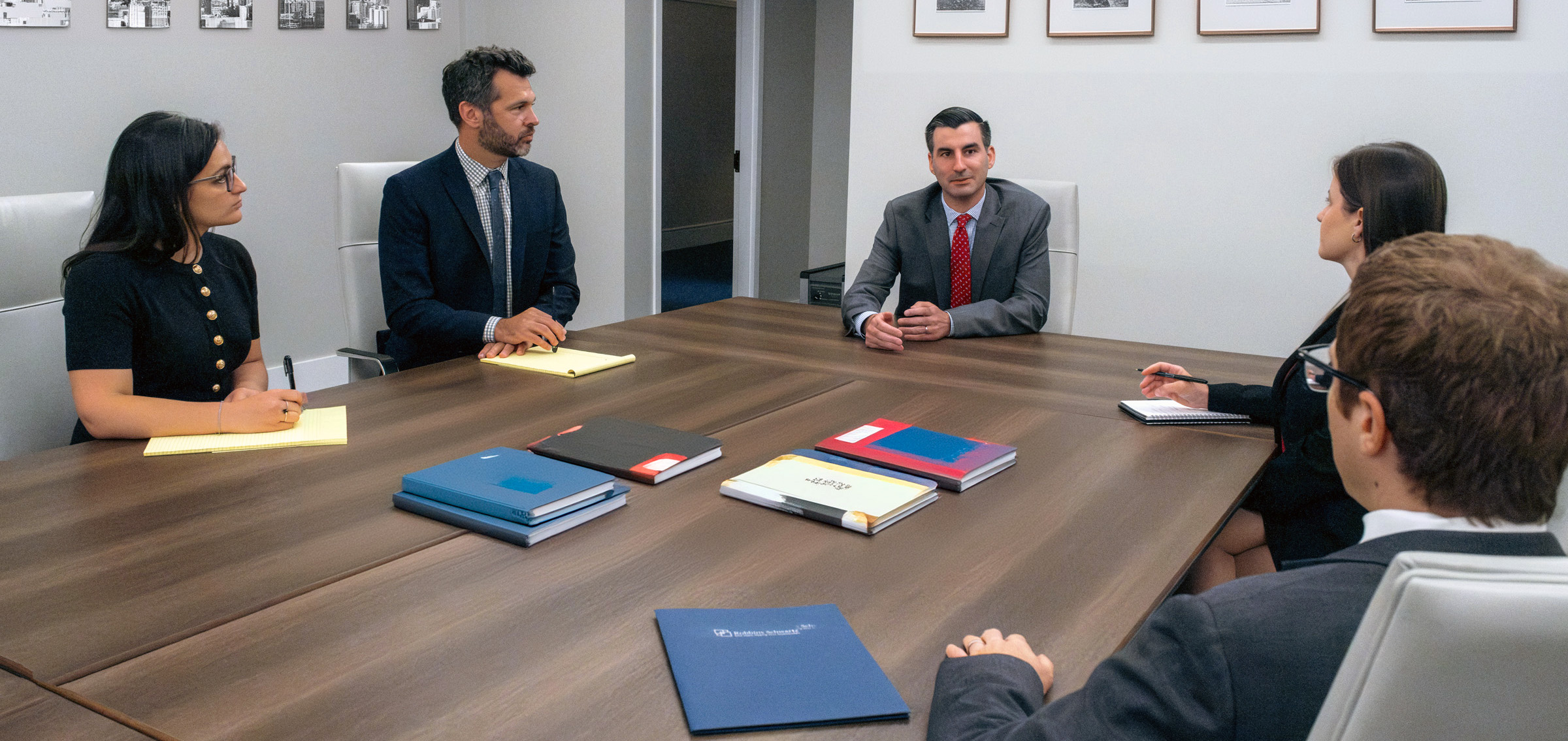 Five business professionals in formal attire seated around a large wooden conference table with folders and documents duri...
