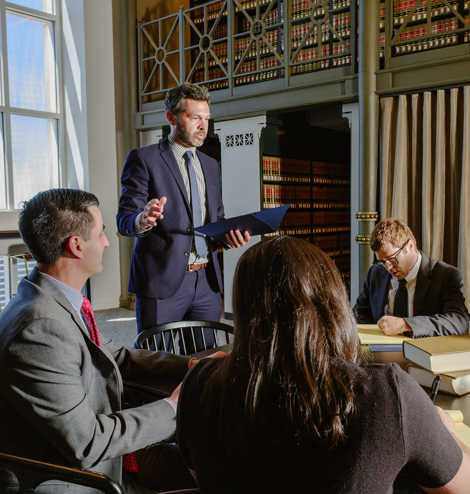 Man in navy suit presenting with a folder to colleagues in a law library lined with legal books during a professional meeting