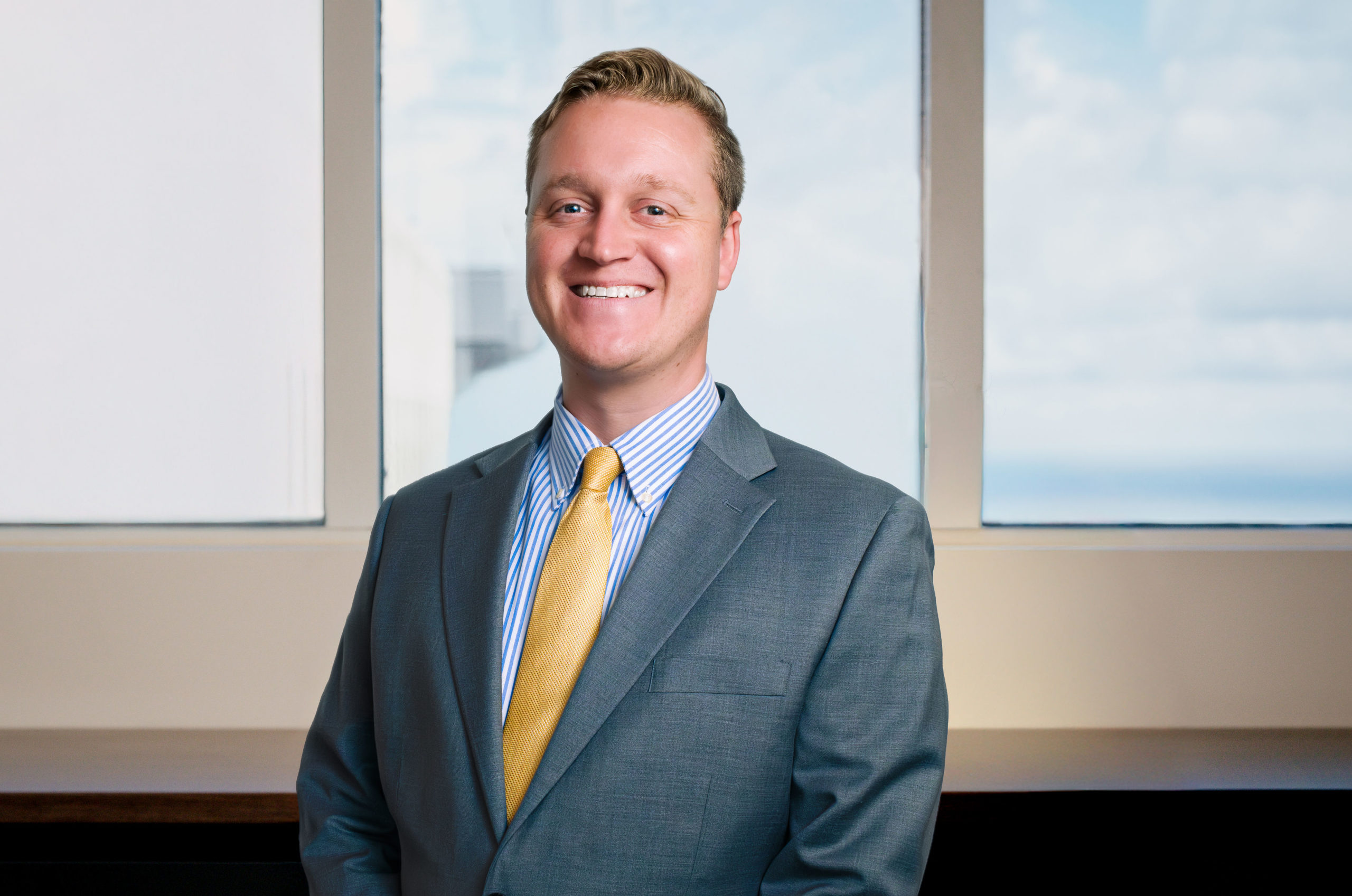 Smiling man in gray suit, blue striped shirt, and gold tie posing in front of large windows with cloudy sky view.