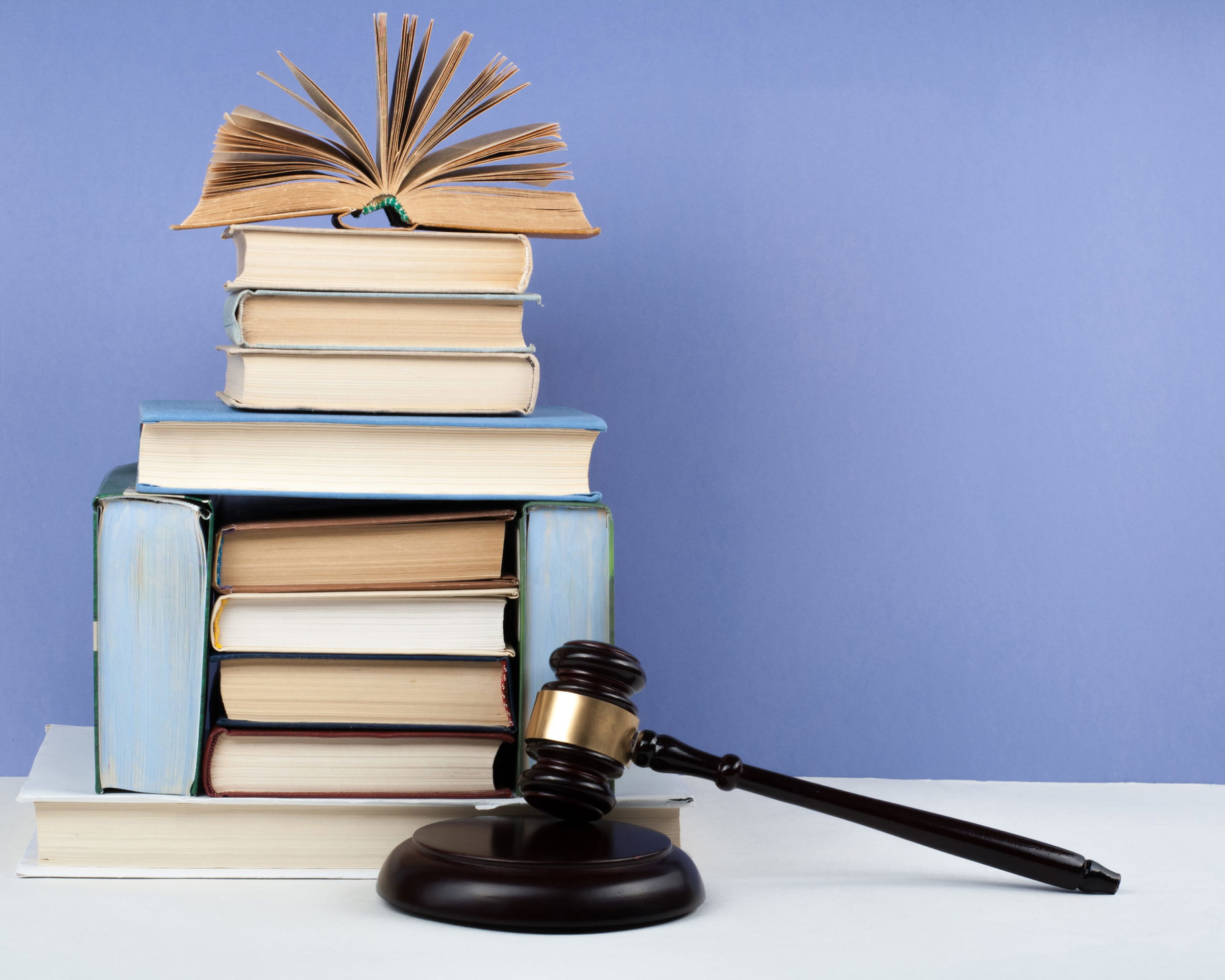 Wooden judge's gavel on a sound block beside a tall stack of law books, with one open book on top, against a blue background.