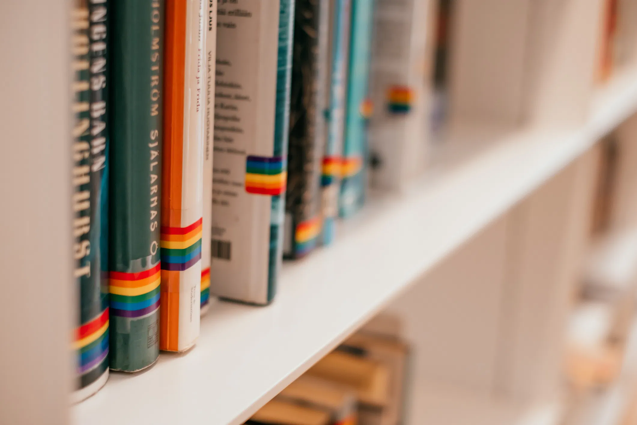 Library shelf with books marked by small rainbow Pride flag stickers on their spines, indicating LGBTQ+ themed or inclusiv...