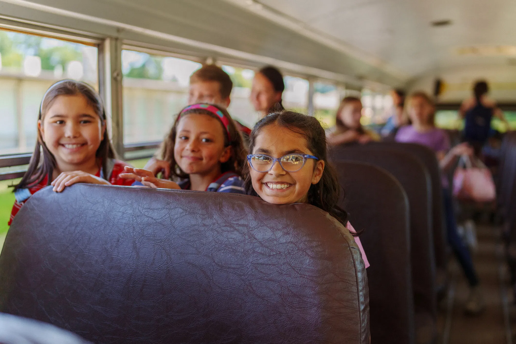 Three smiling elementary school girls riding a yellow school bus, with other students seated in the background.