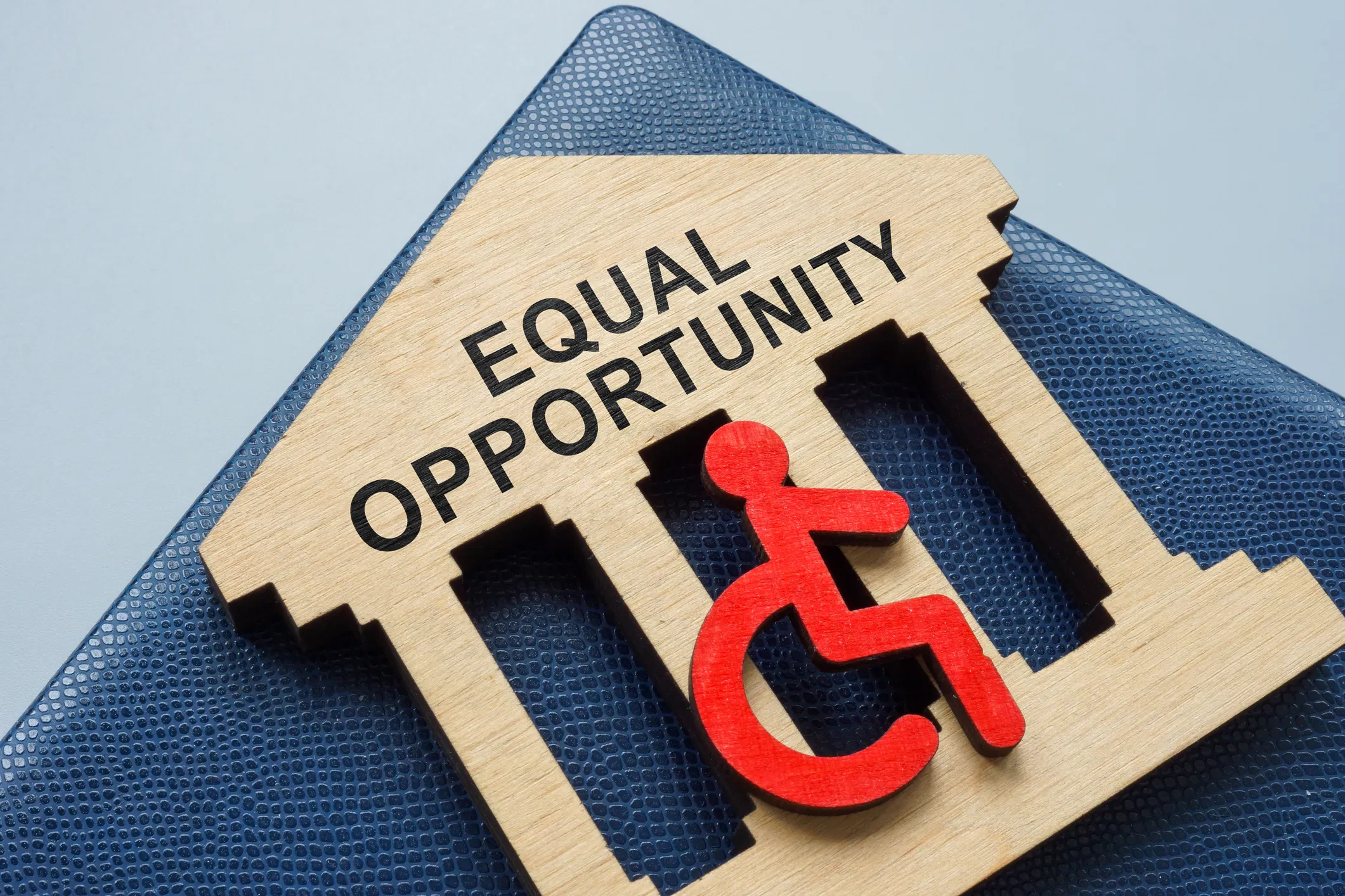 Wooden building cutout reading "Equal Opportunity" with a red wheelchair accessibility symbol, resting on a blue textured ...