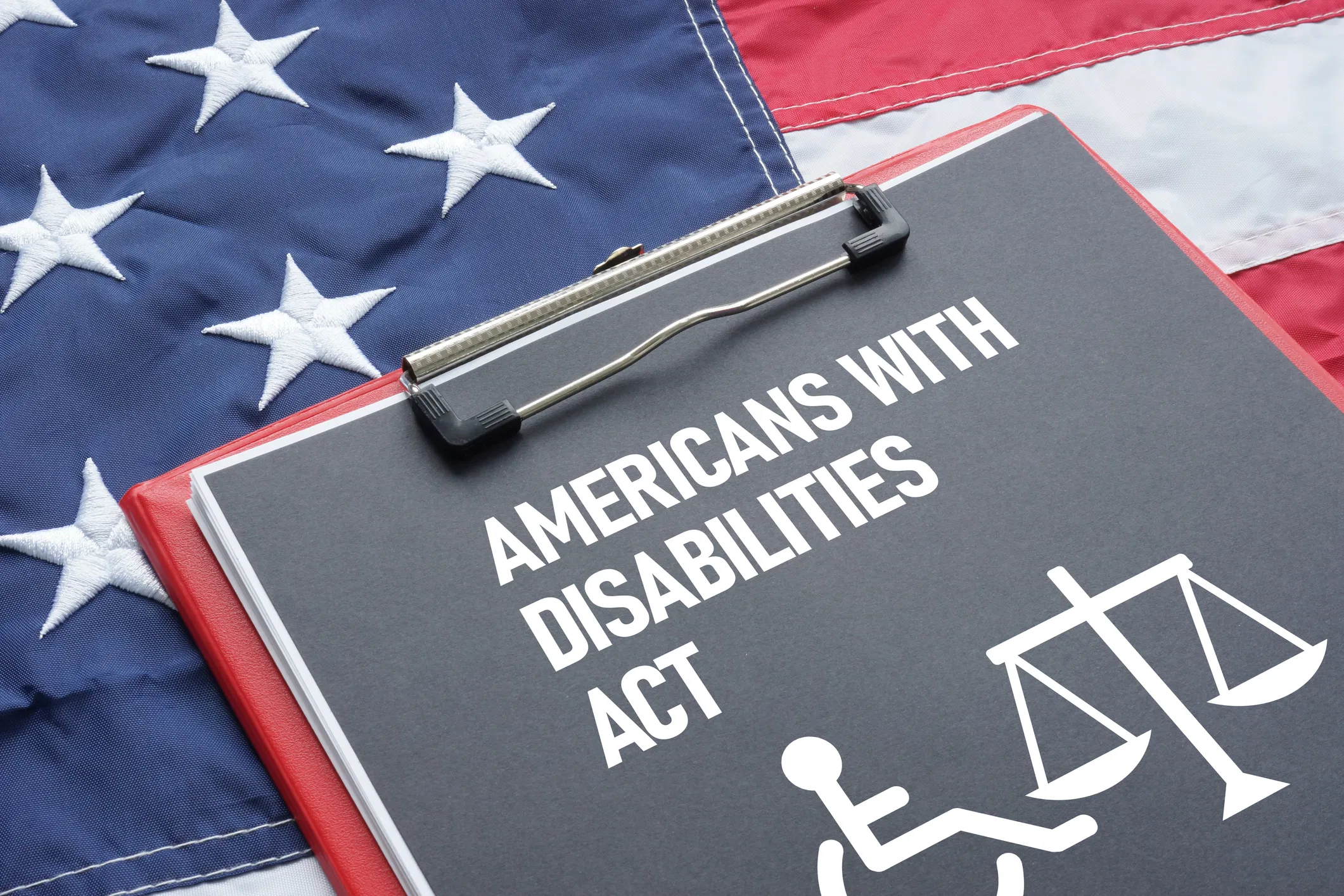 Clipboard reading "Americans With Disabilities Act" with wheelchair and scales of justice icons, resting on a US flag