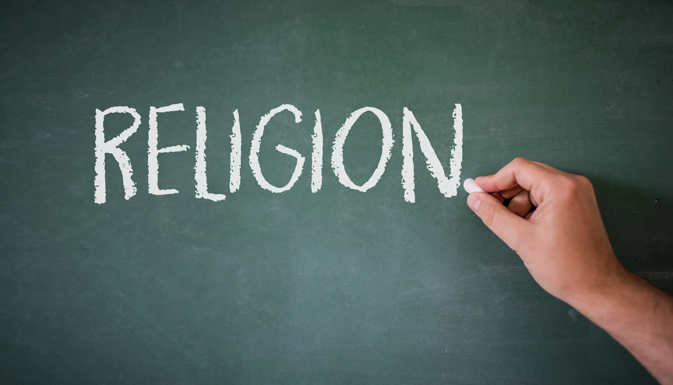 Hand writing the word RELIGION in white chalk on a green chalkboard