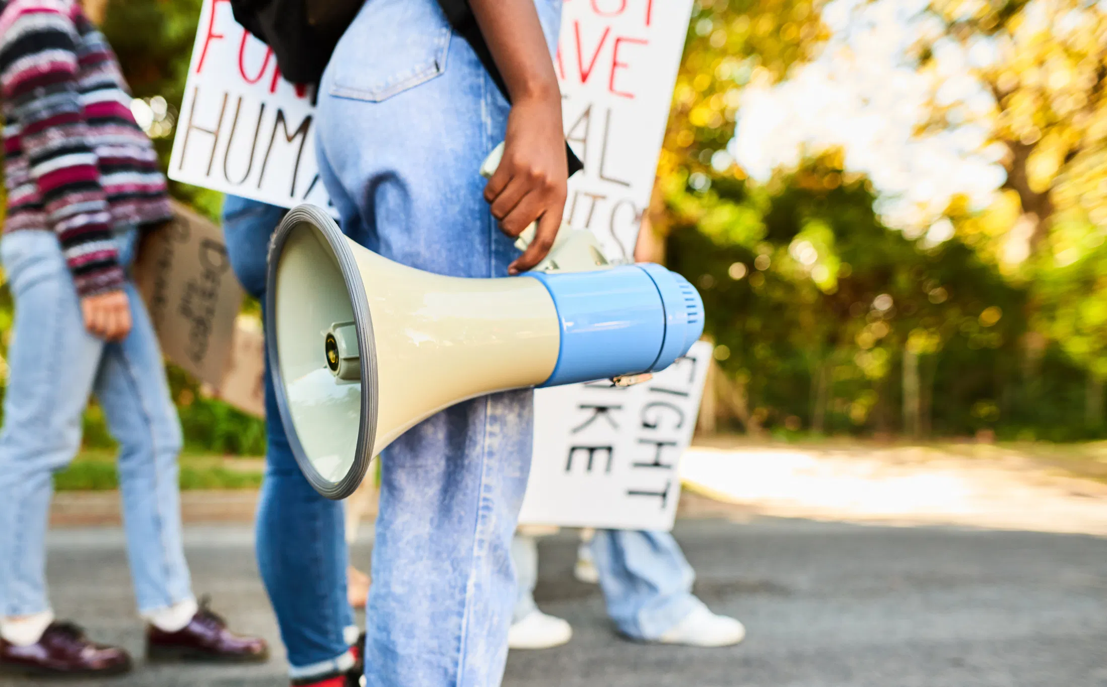 Person holding a blue and cream megaphone at an outdoor protest, with demonstrators carrying human rights signs in the bac...