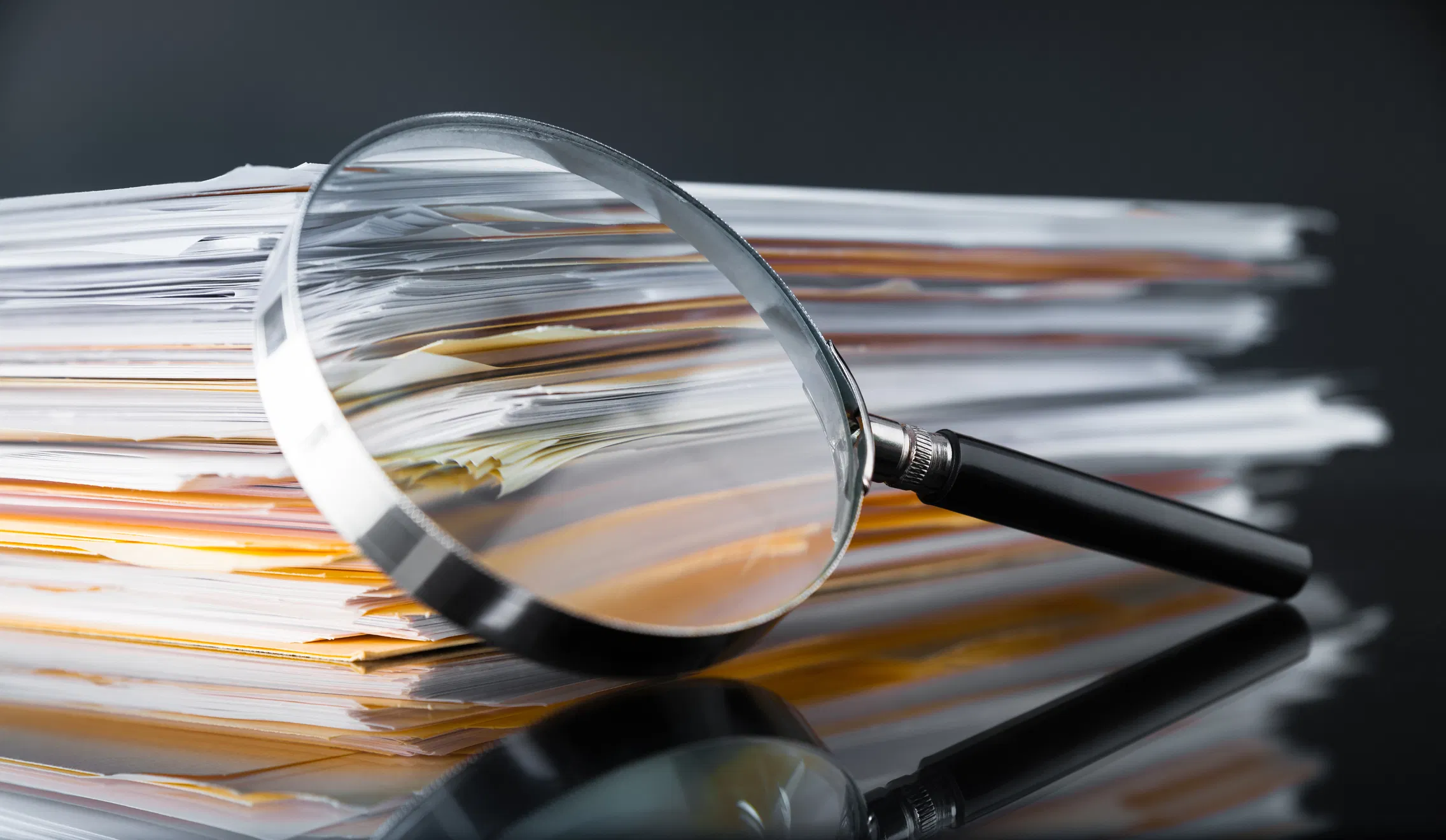 Magnifying glass leaning against a large stack of white and yellow paper documents on a reflective dark surface