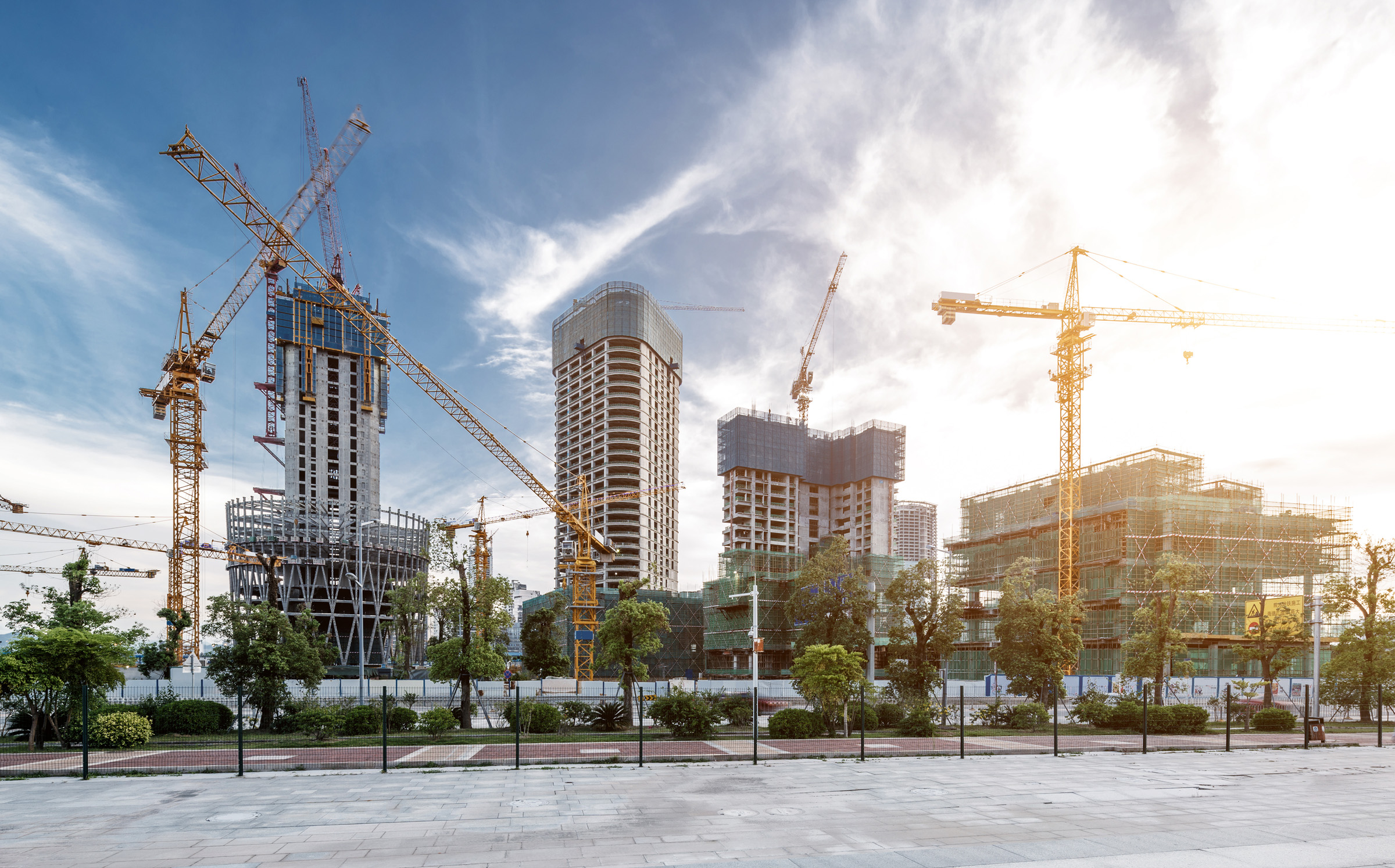 Multiple high-rise buildings under construction with yellow tower cranes against a partly cloudy sky, viewed from a paved ...