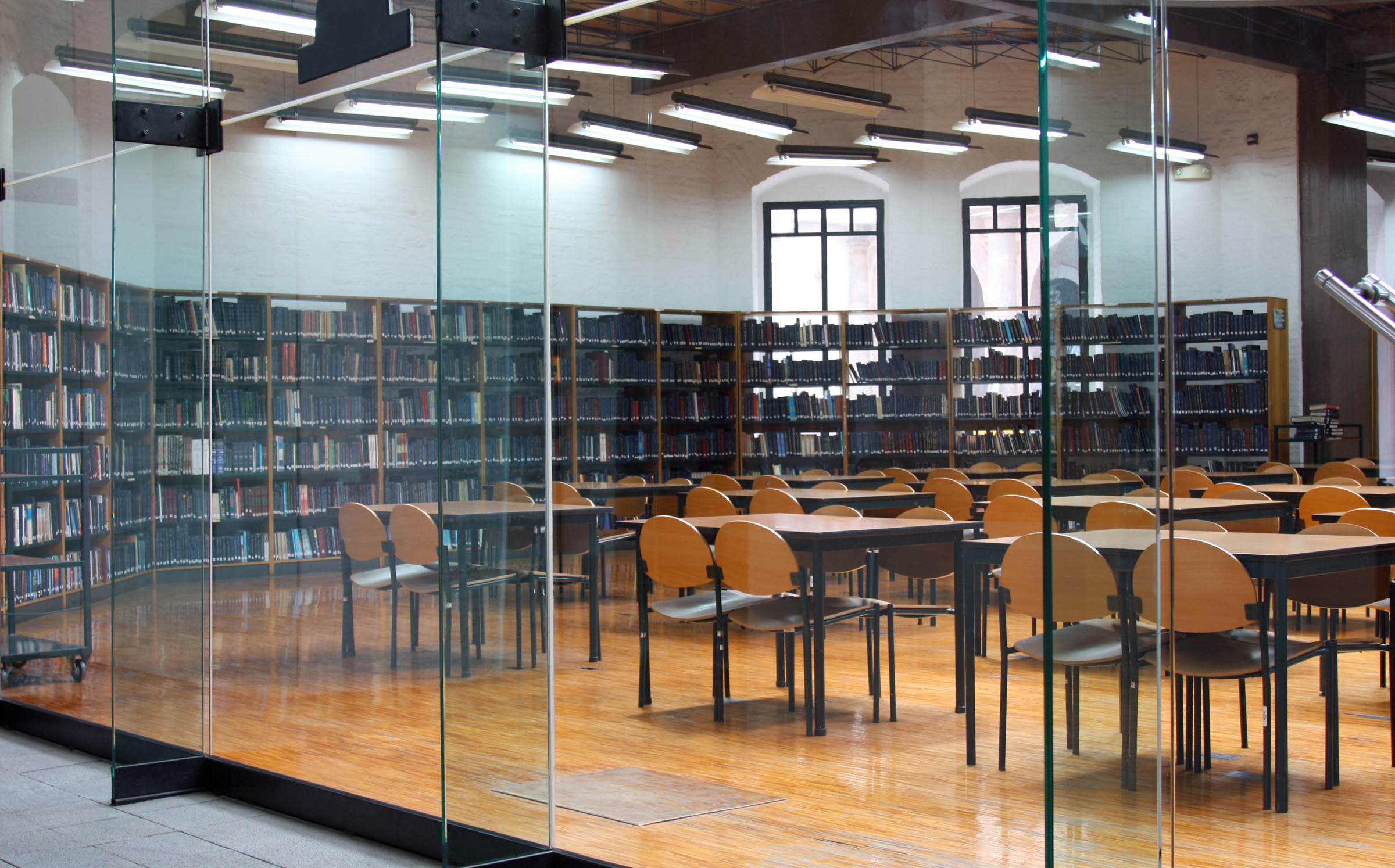 Empty library reading room with wooden chairs and tables, floor-to-ceiling glass walls, and tall bookshelves lining the wa...