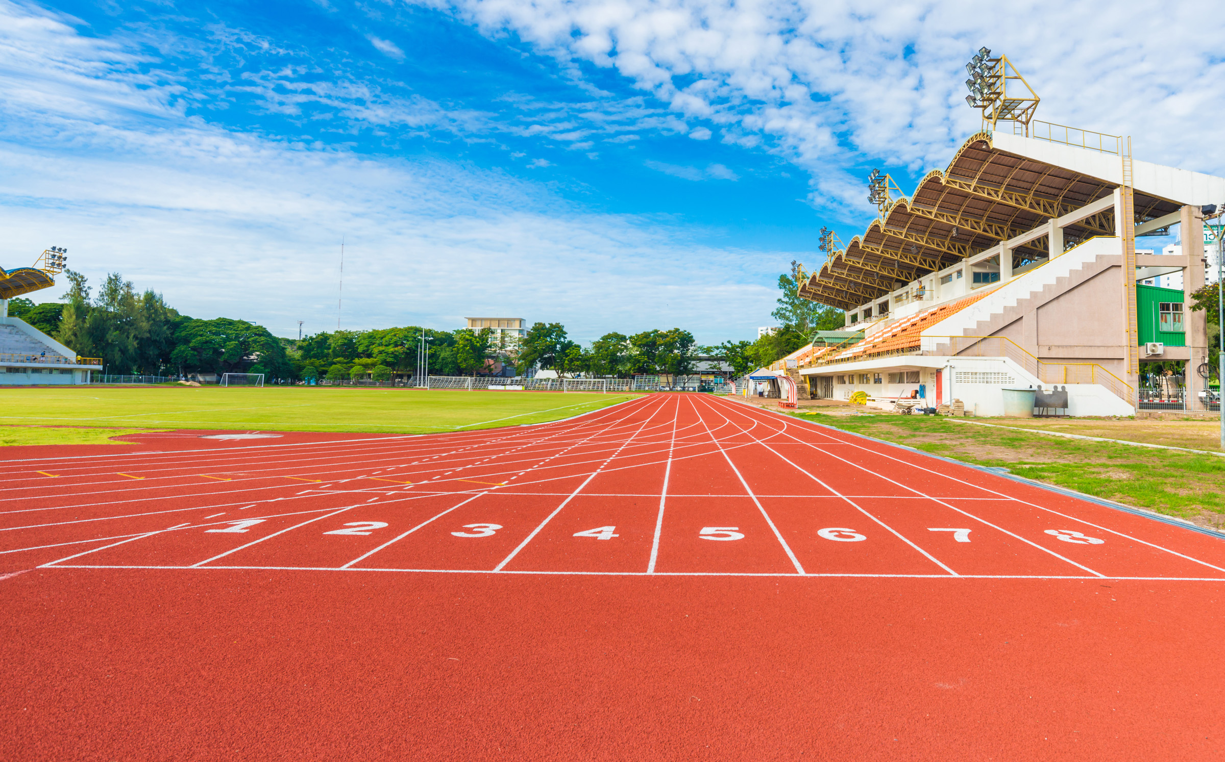 Empty red running track with 8 numbered lanes at an outdoor stadium with grandstands, green infield, and blue sky
