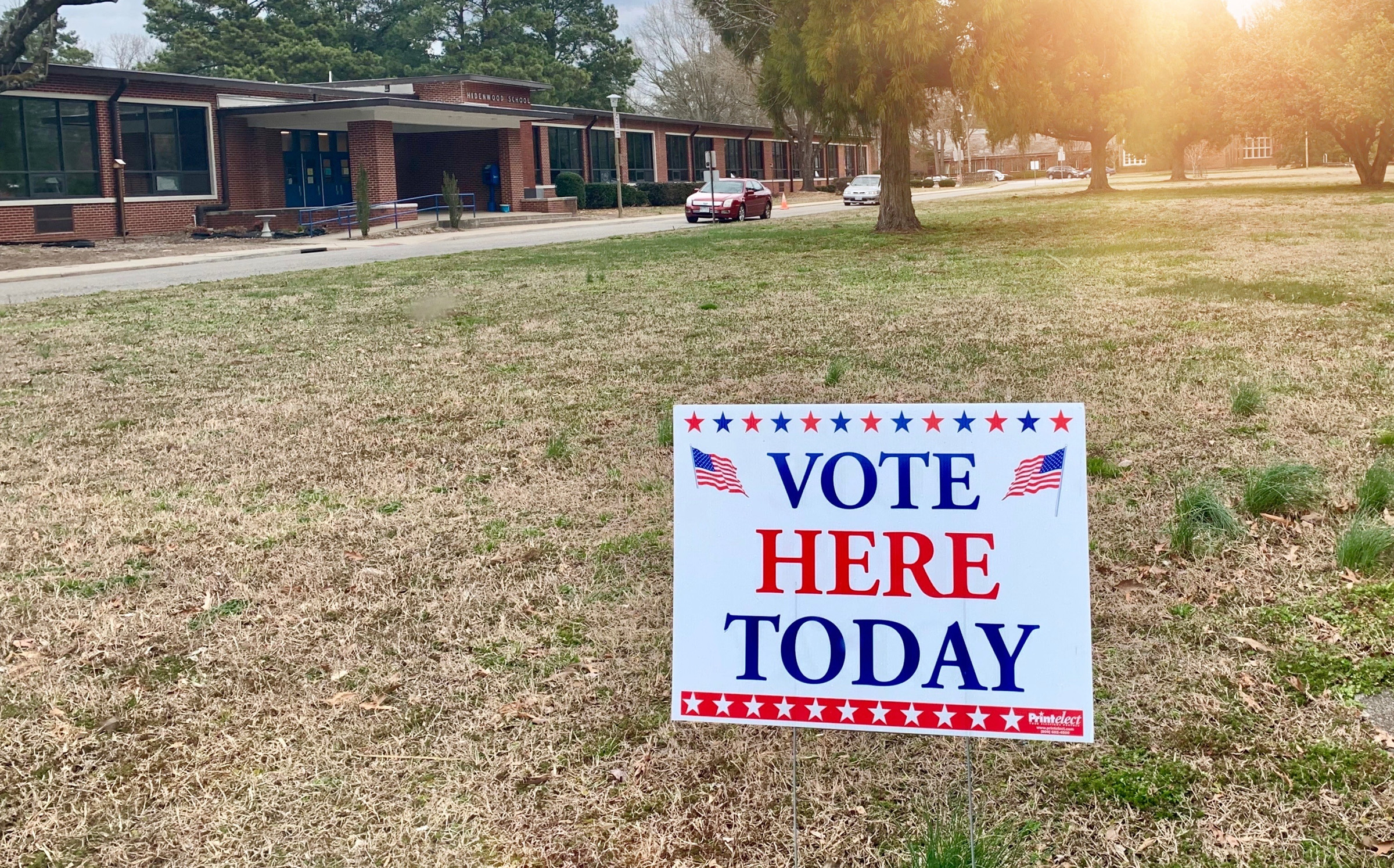 Vote Here Today yard sign with American flags and stars on dry grass outside a brick polling place building
