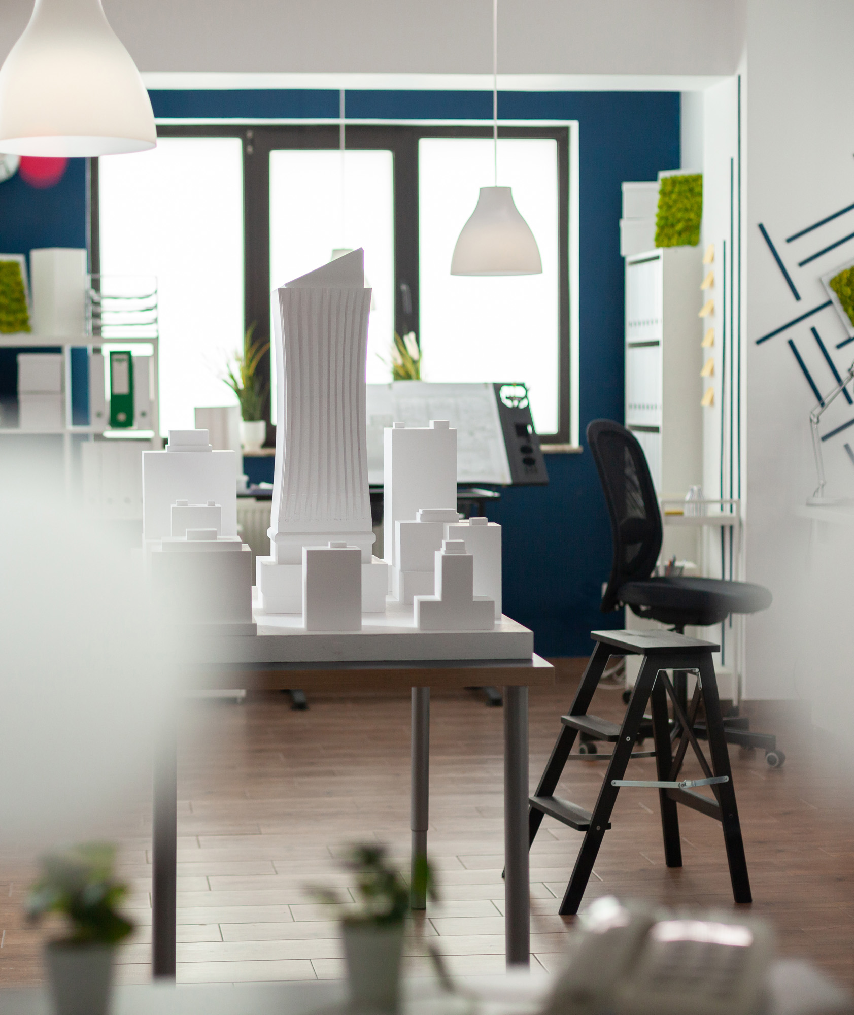 White architectural scale model of city buildings on a desk in a modern architecture studio with blue accent walls and pen...