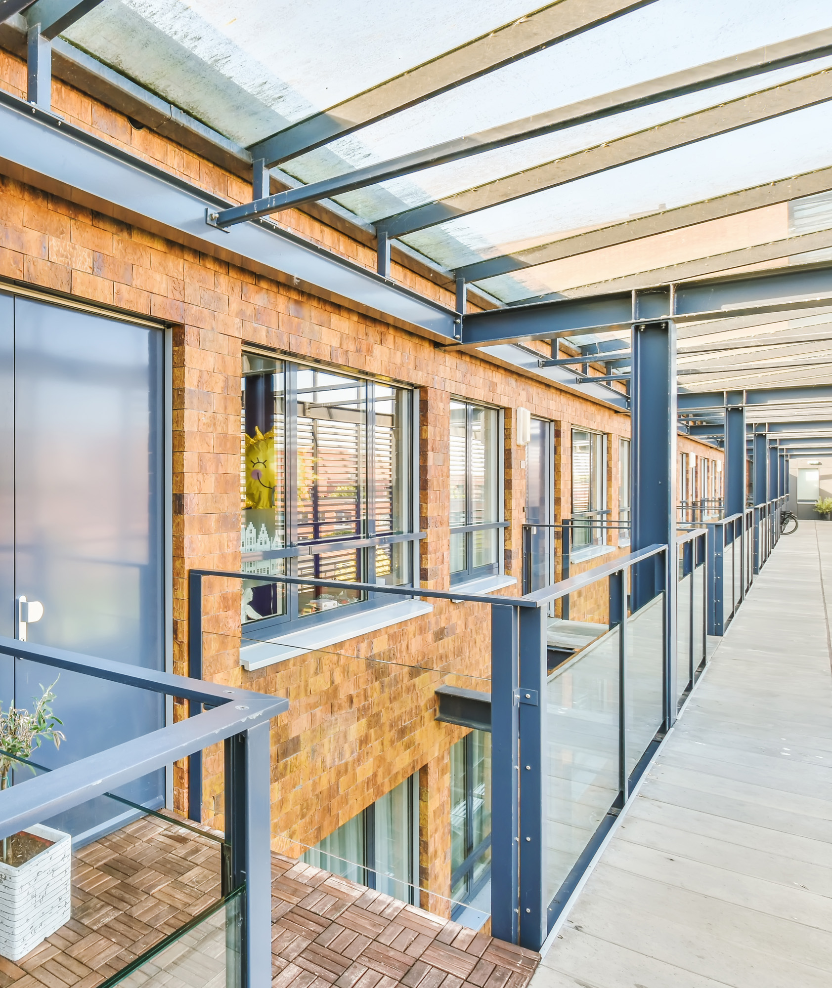 Covered outdoor corridor of a modern brick building with blue steel frame, glass railings, and large windows along upper-f...