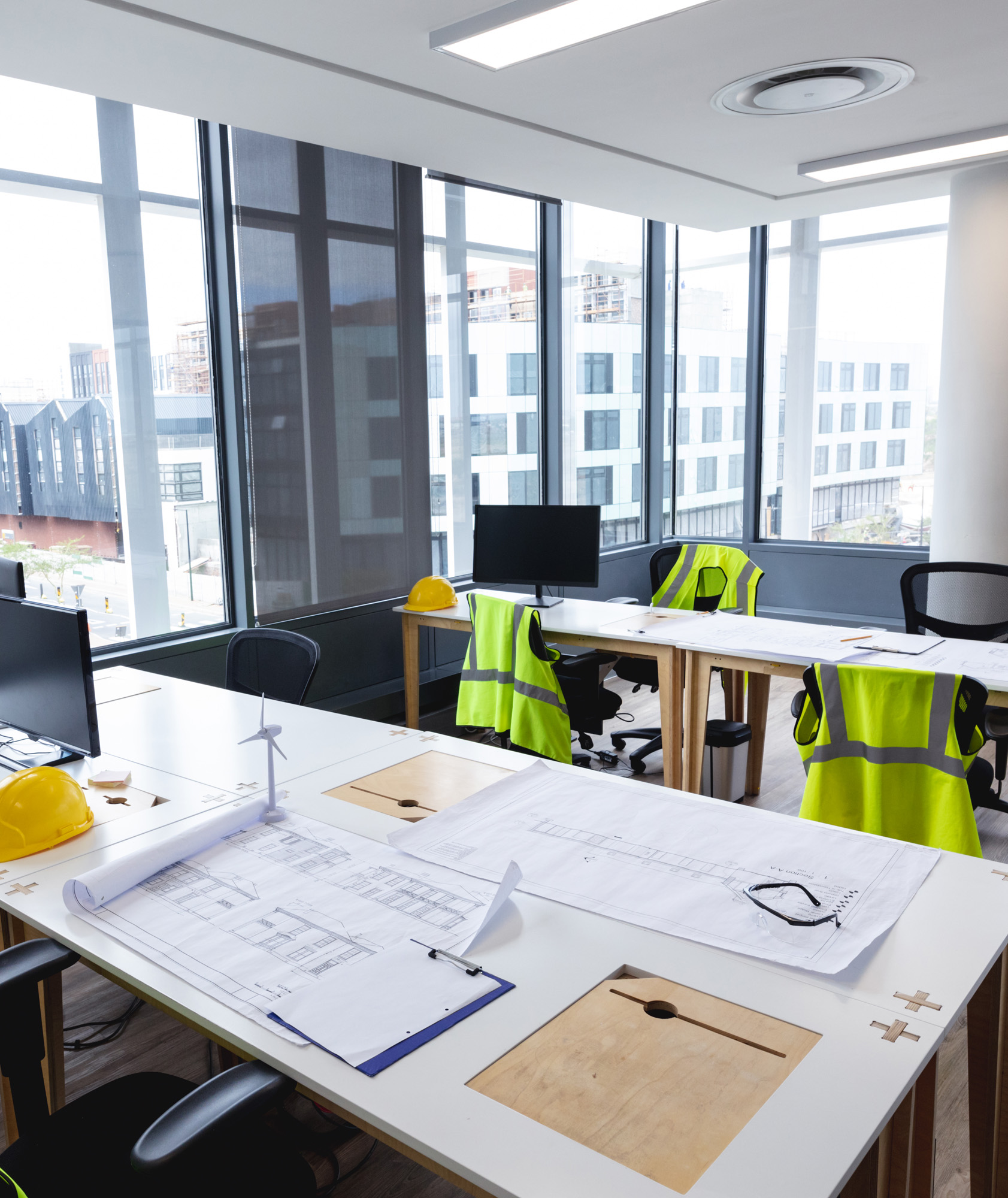Construction office workspace with blueprints, yellow hard hat, wind turbine model, and hi-vis vests on chairs, overlookin...