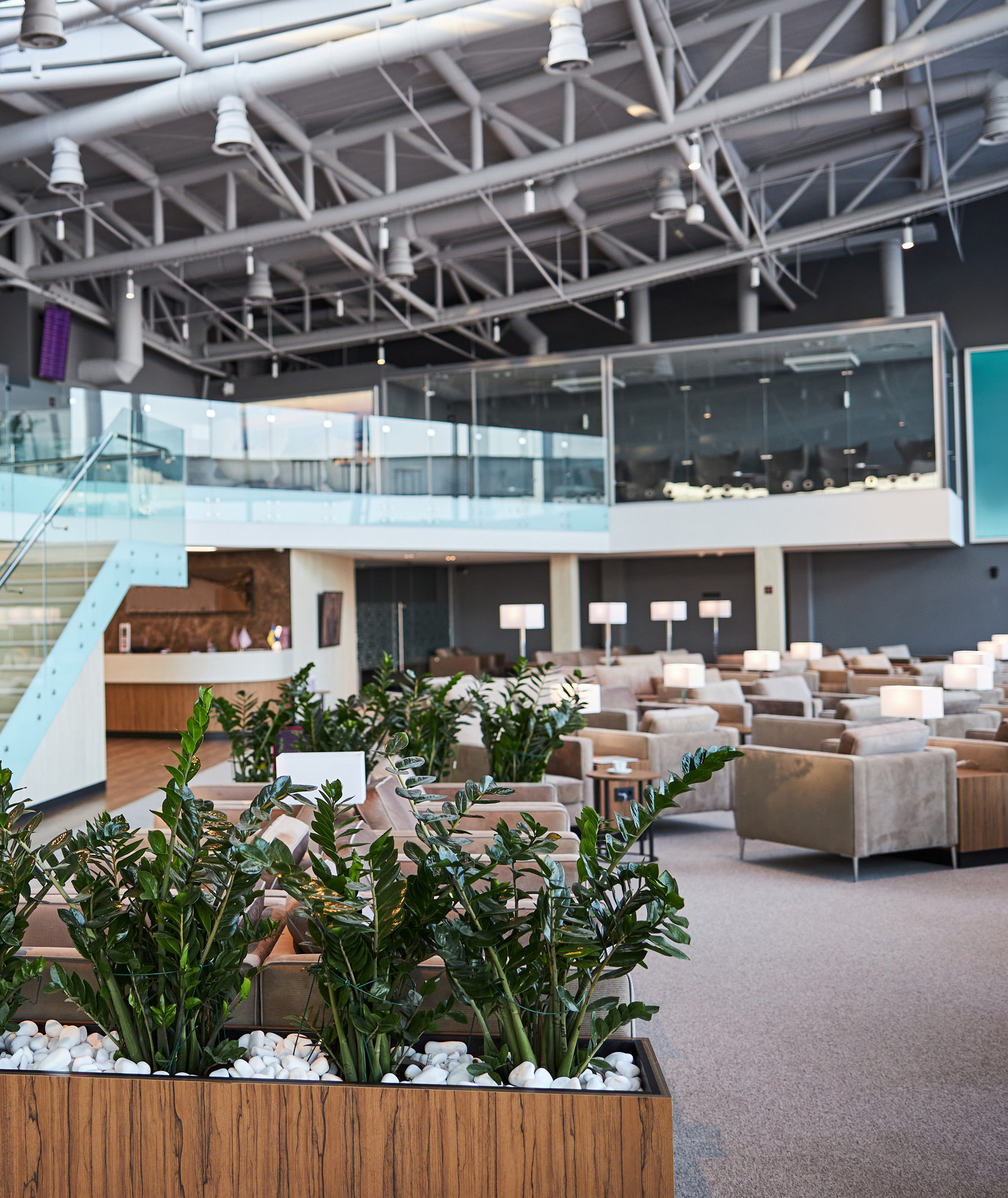Modern airport lounge with ZZ plants in wooden planters, beige seating areas with table lamps, glass mezzanine level, and ...