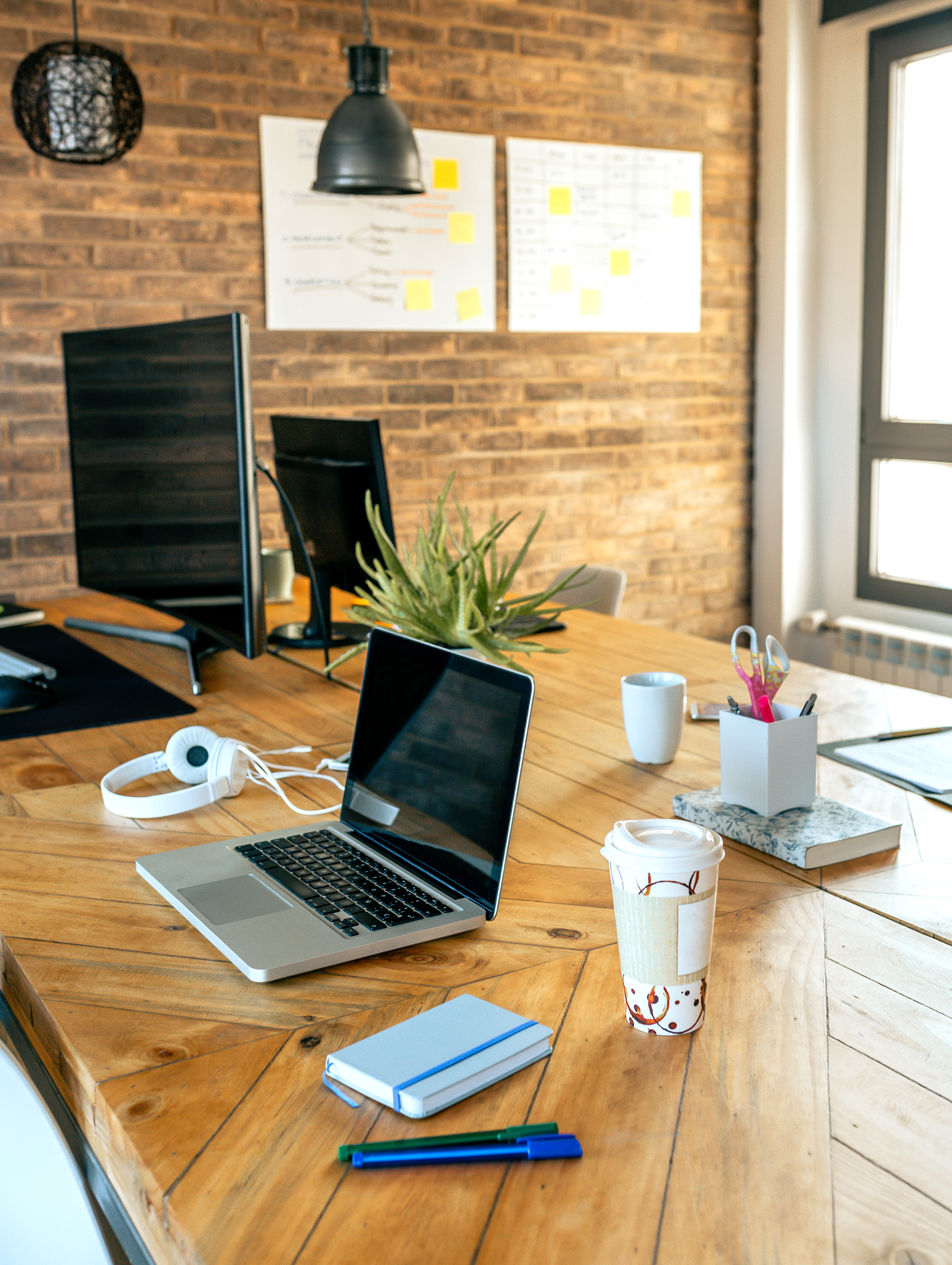 Modern workspace with open laptop, takeaway coffee cup, headphones, notebook, and pens on a wooden desk against an exposed...