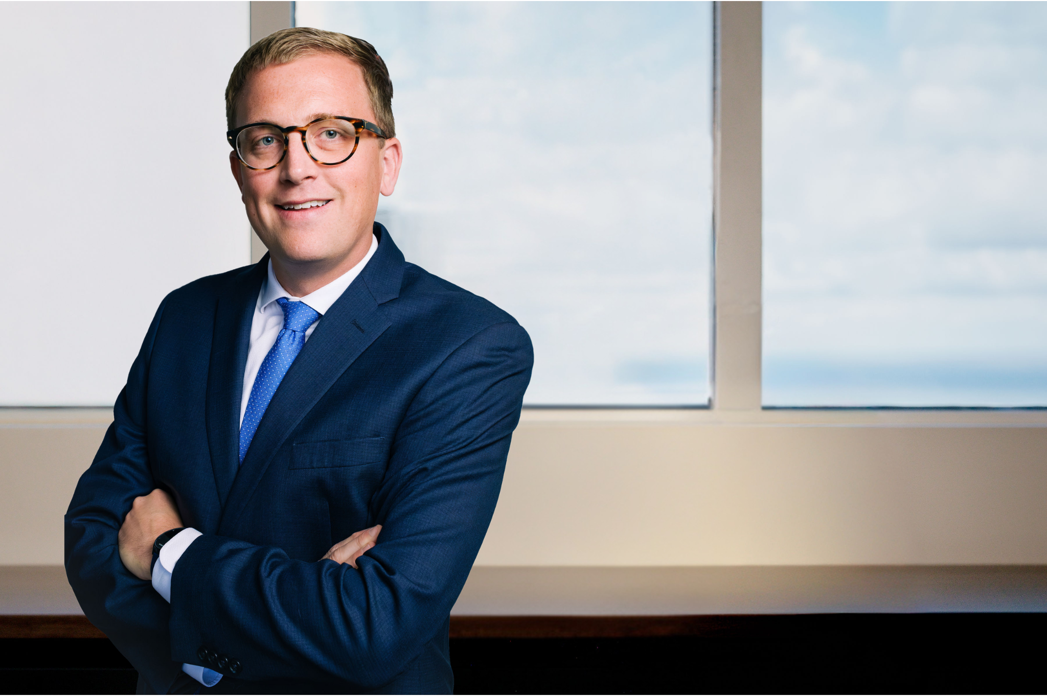 Man in navy suit and blue tie with arms crossed, smiling, wearing glasses, standing before a window with ocean view