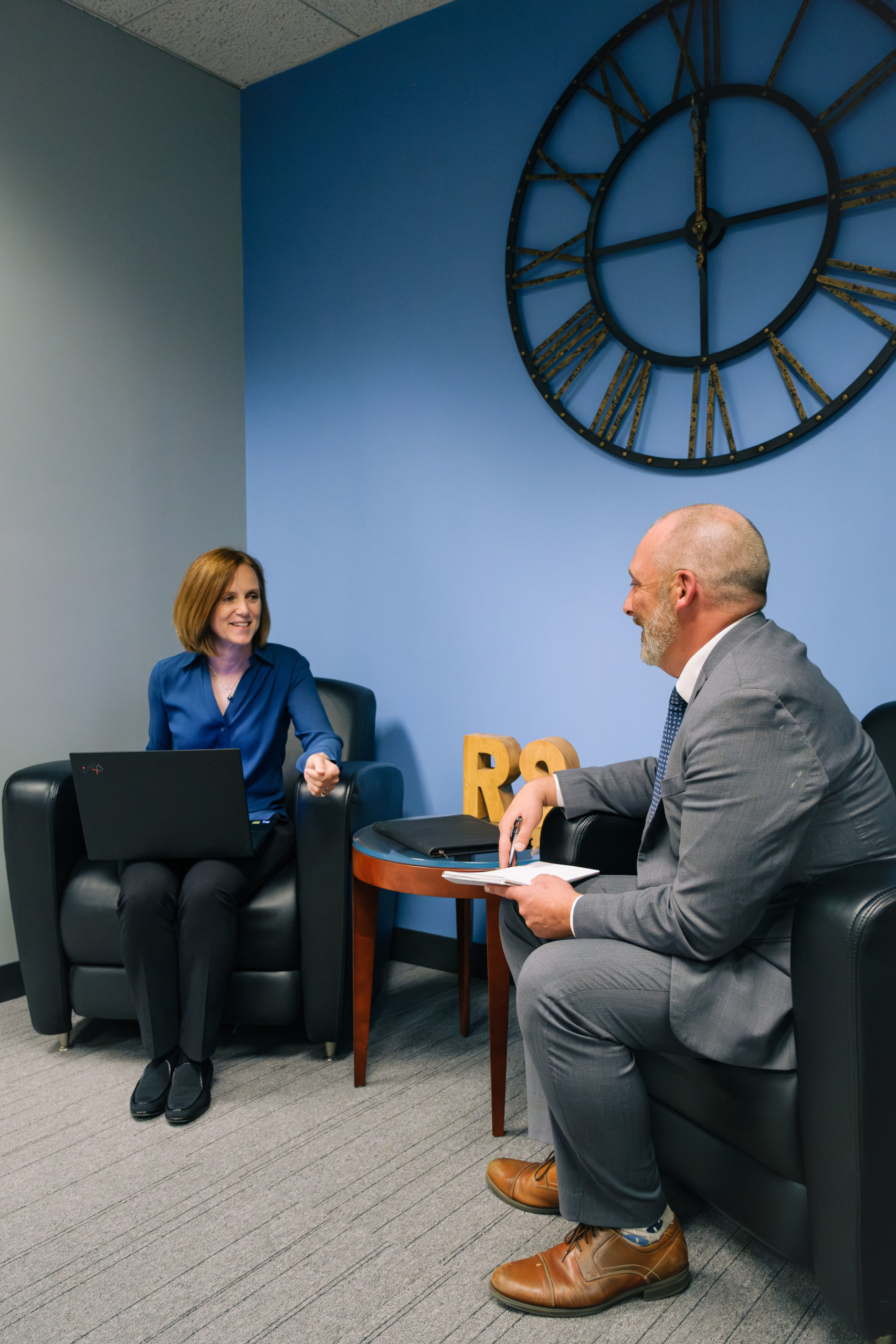Two professionals in a business meeting, woman with laptop in blue blouse facing man in gray suit holding notepad, large c...
