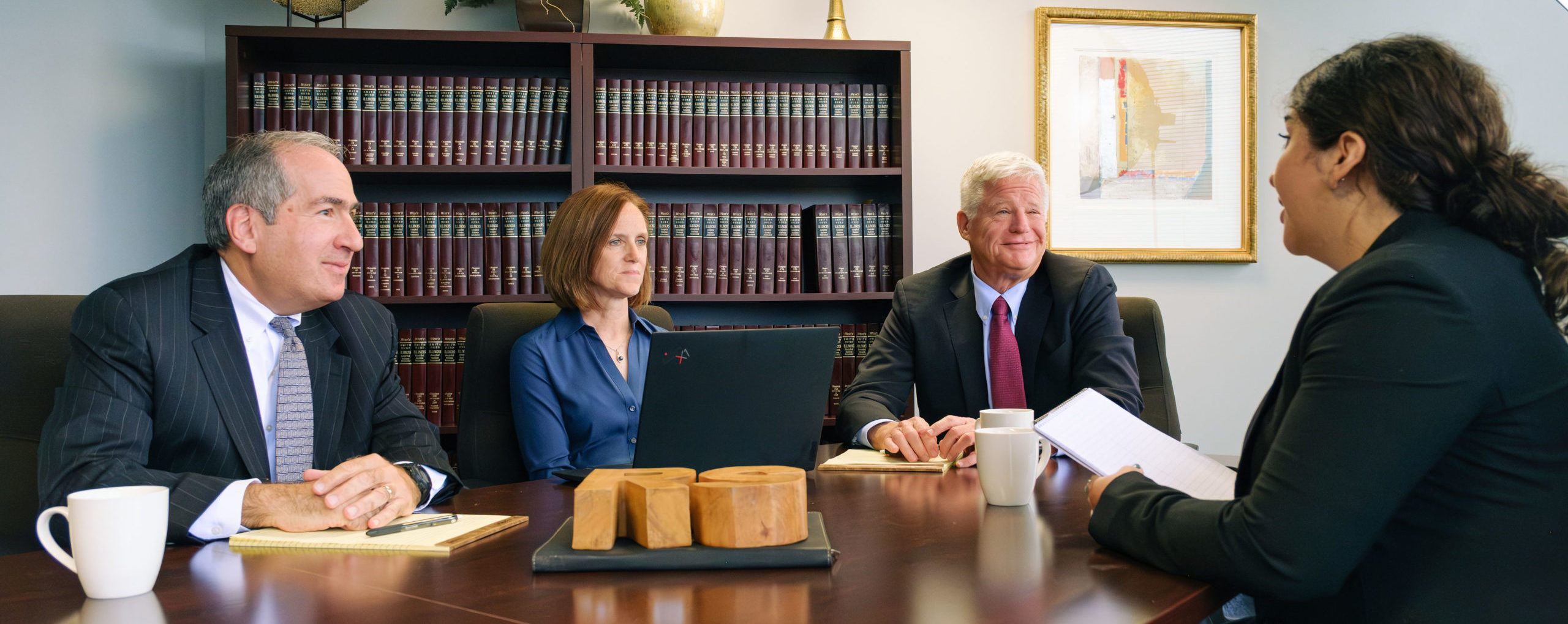 Four professionals in business attire meeting around a conference table in a law office with floor-to-ceiling bookshelves ...