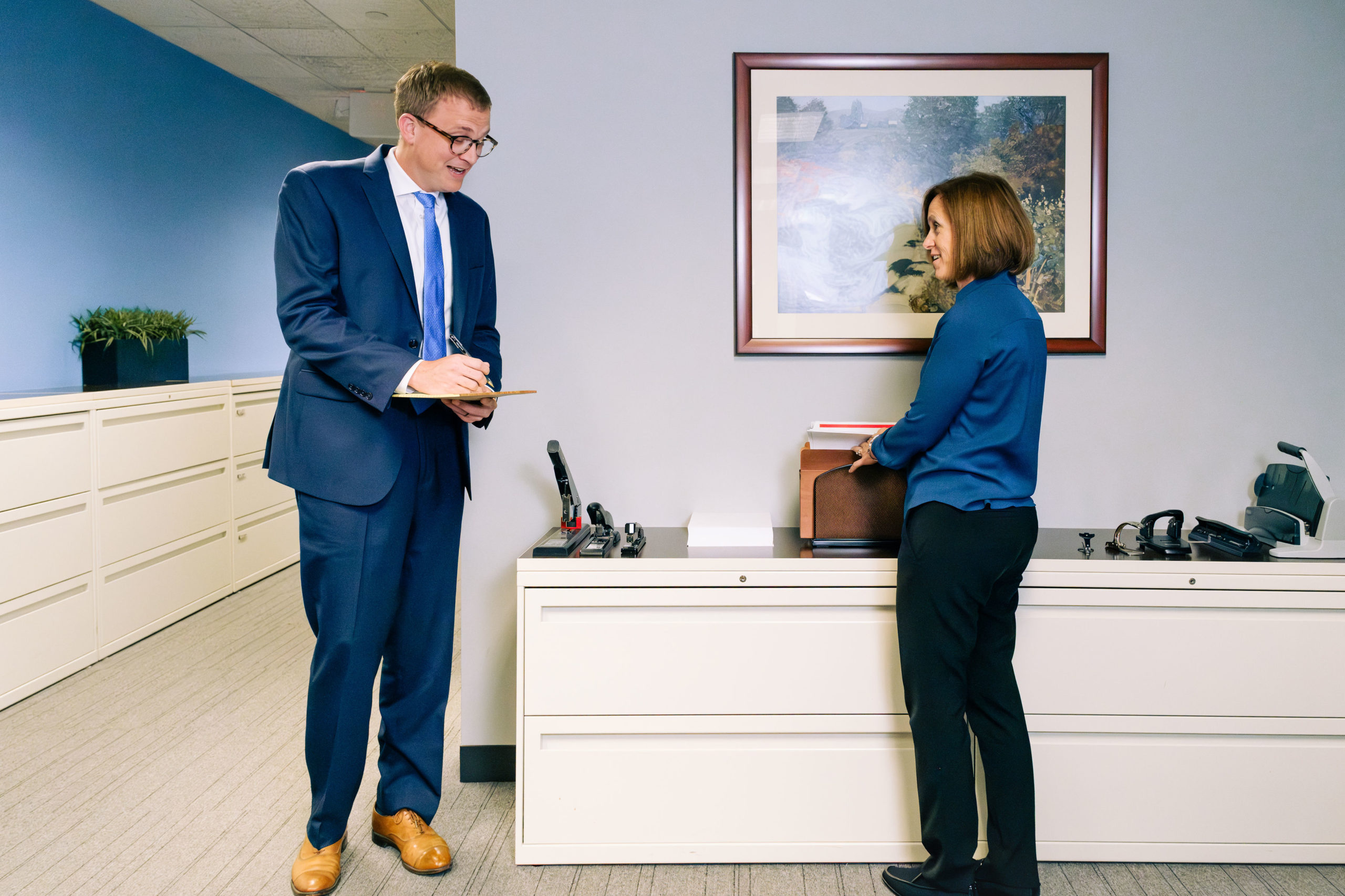 Man in navy suit holding clipboard speaks with woman in blue top leaning against office filing cabinet beneath a framed ar...
