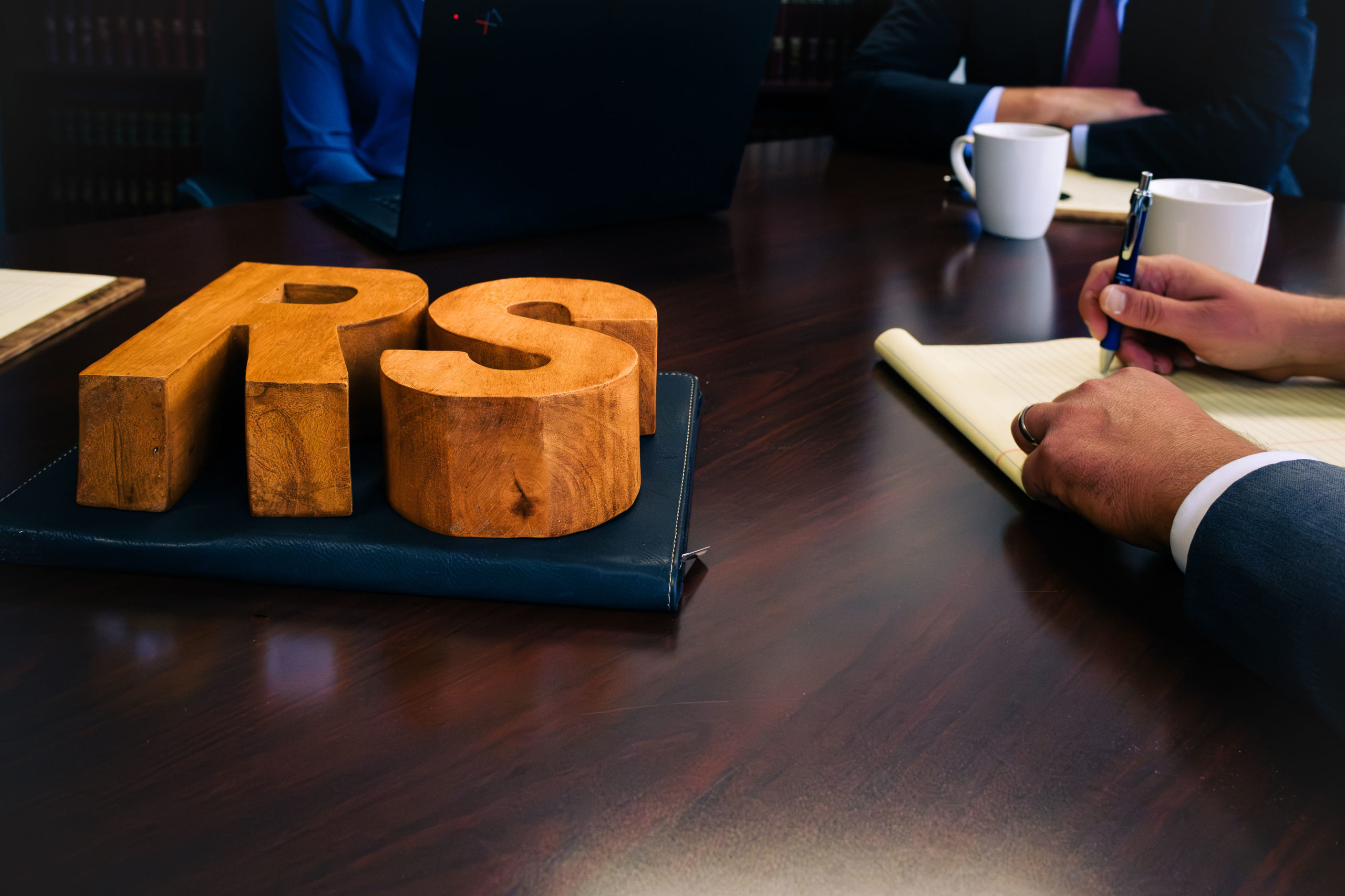 Wooden letters "RS" on a leather folder at a conference table where suited professionals take notes and use a laptop with ...