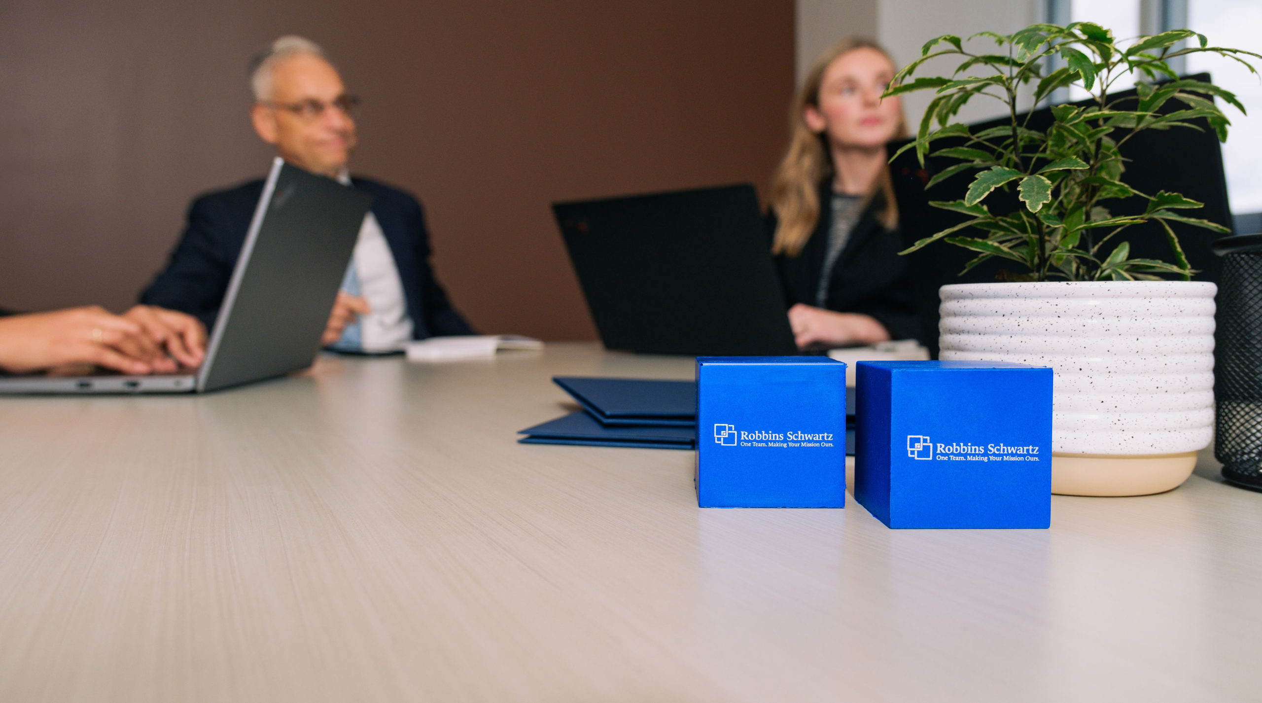 Two blue branded gift boxes labeled Robbins Schwartz on a conference table, with a potted plant and blurred woman on lapto...
