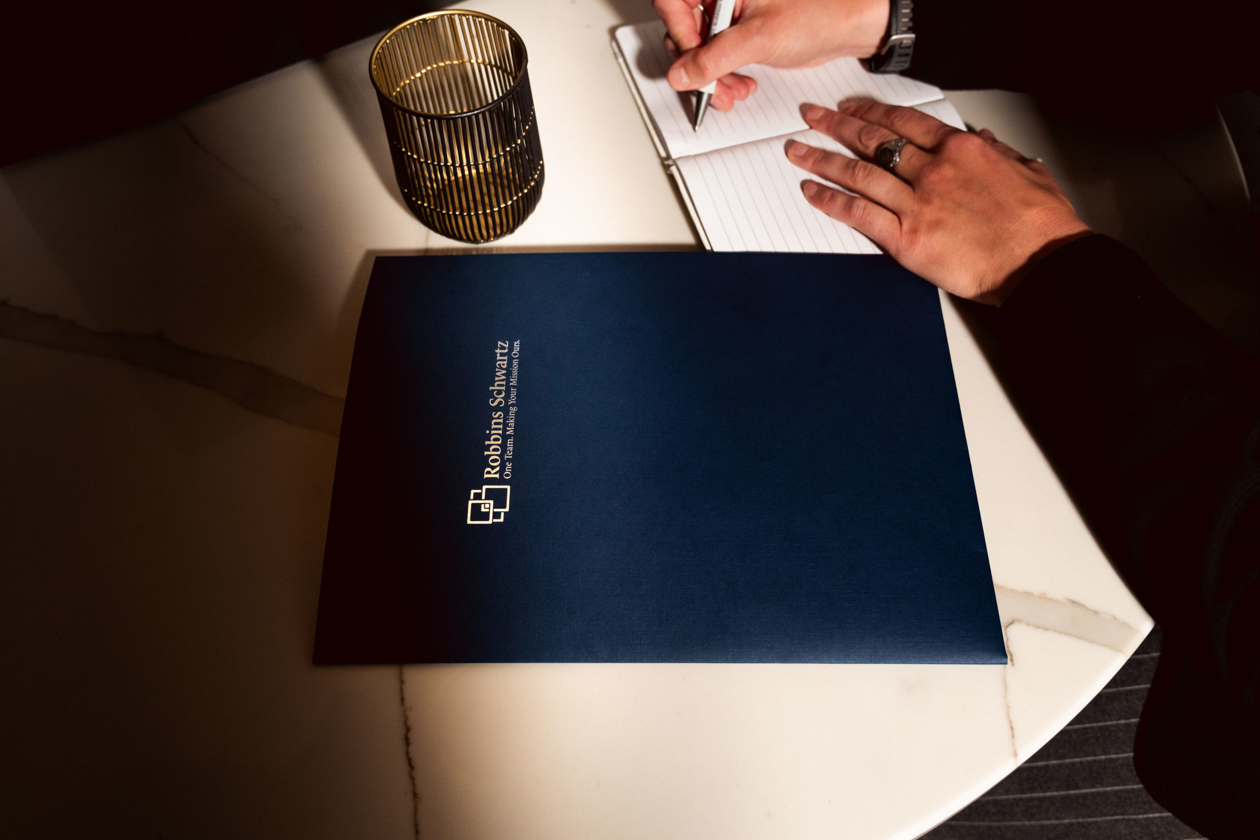 Navy blue folder with Robbins Schwartz logo and tagline on a white table, with a person's hand and notepad nearby.