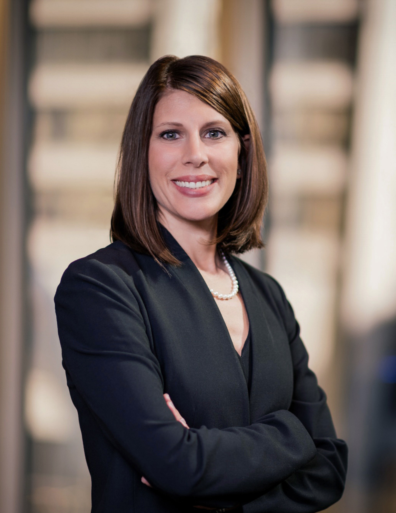 Woman with brown hair in black blazer and pearl necklace smiling with arms crossed in a professional headshot