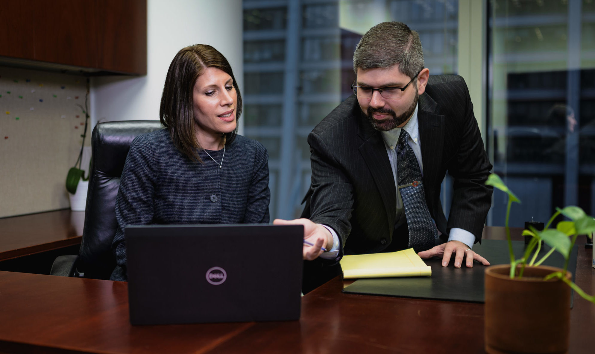 Two business professionals in suits reviewing content on a Dell laptop at an office desk, man pointing at screen while hol...