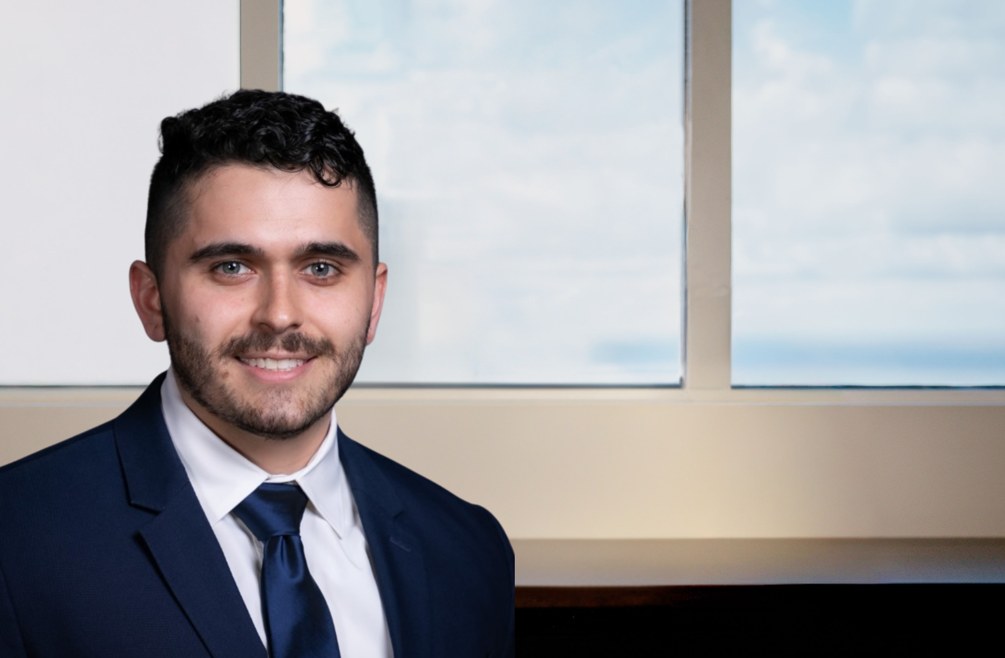Young man with dark curly hair and beard smiling, wearing a navy suit and blue tie, seated near a window with sky view.
