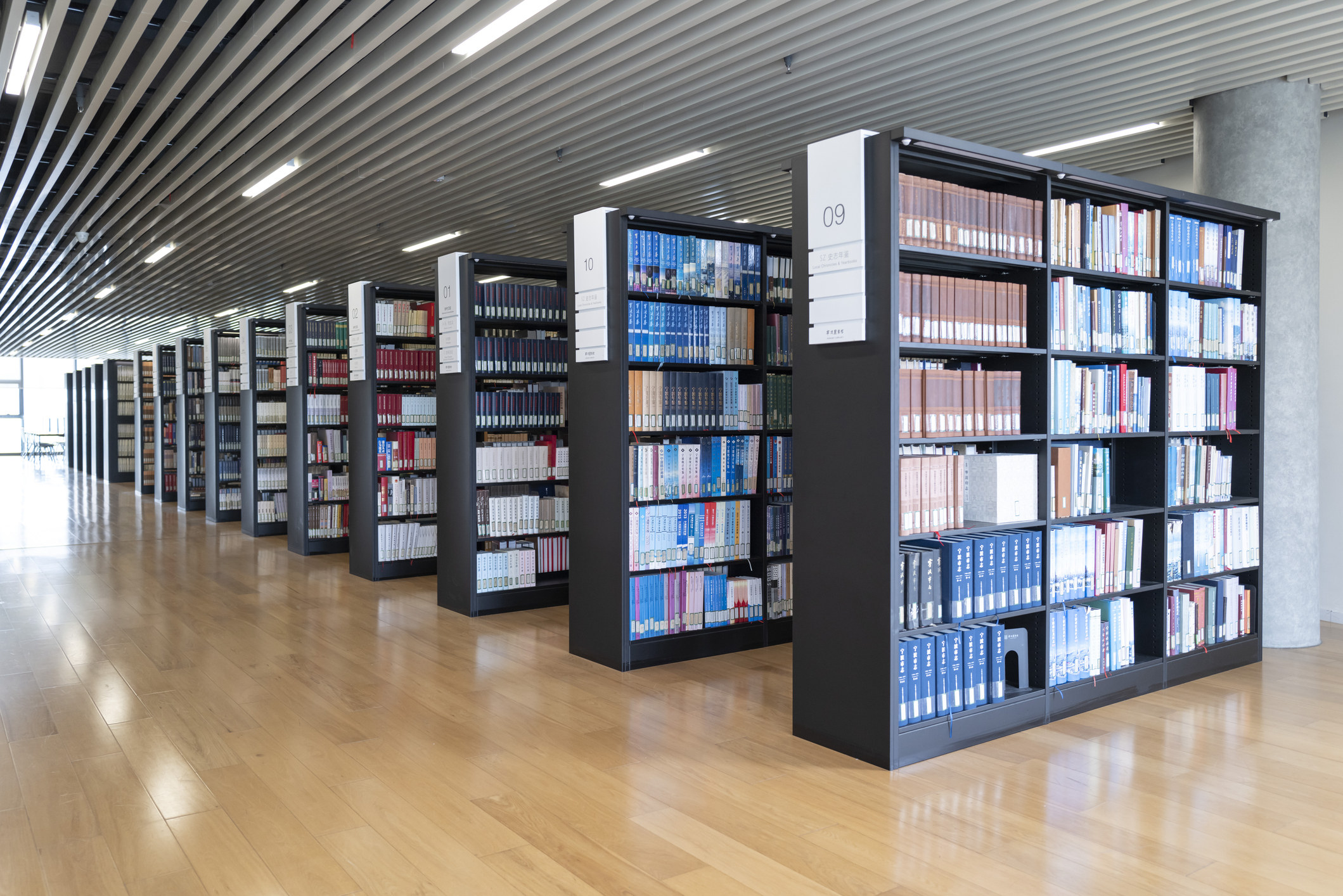 Rows of dark metal library shelves filled with colorful books on polished wood floors under linear ceiling lights, labeled...