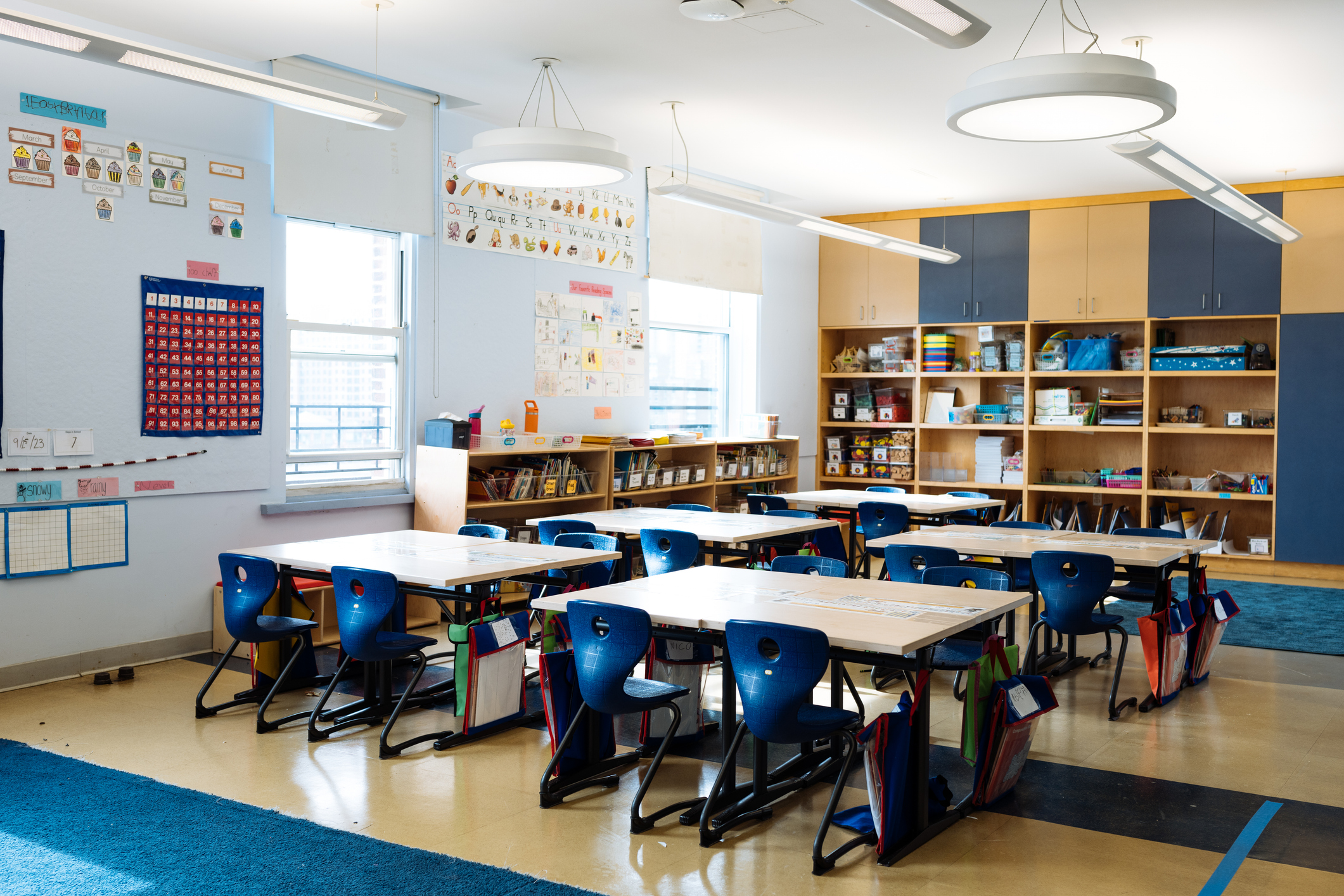 Empty elementary school classroom with blue chairs, grouped desks, wooden bookshelves, and colorful educational posters on...