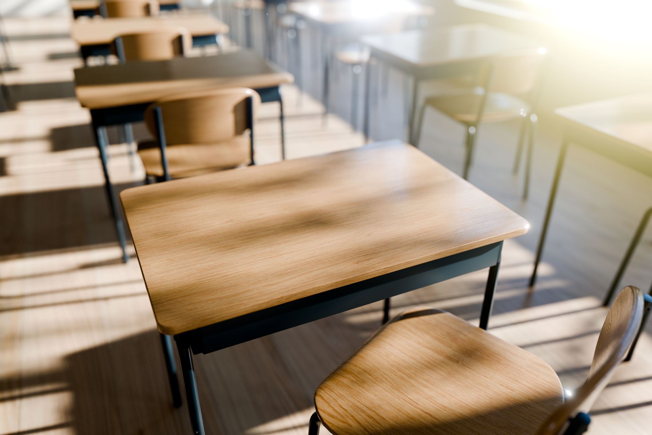 Empty classroom with rows of wooden student desks and chairs on a light wood floor, sunlight streaming through windows