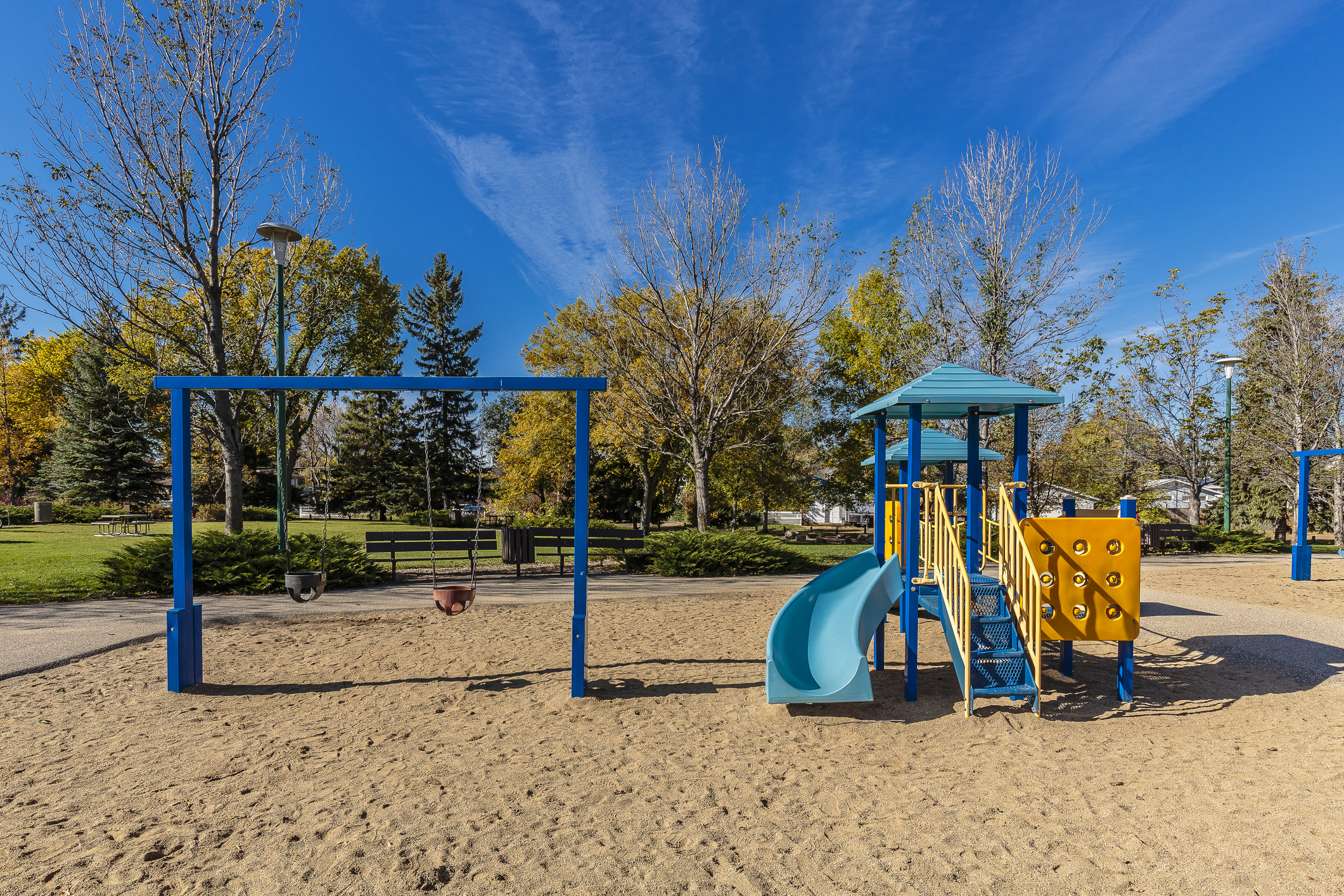 Empty playground with blue and yellow play structure, light blue slide, and blue swing set on sandy ground surrounded by t...