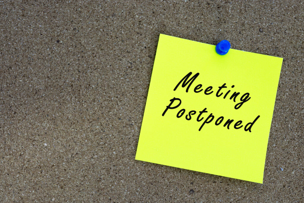 Meeting postponed on yellow stick note and pinned to a cork notice board. Reminder and business concept.