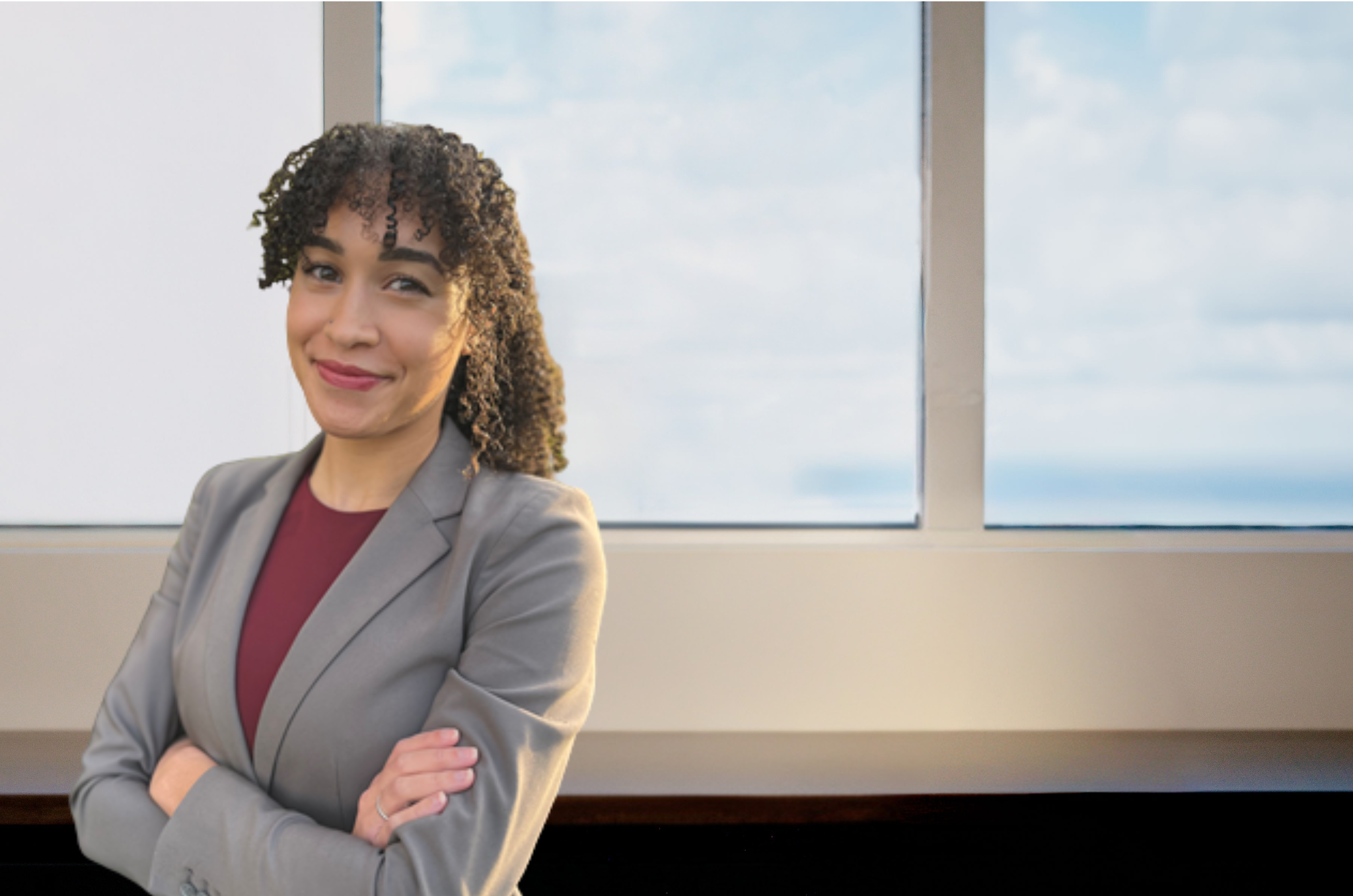 Woman with curly hair in a gray blazer and burgundy top standing arms crossed, smiling confidently near large office windows