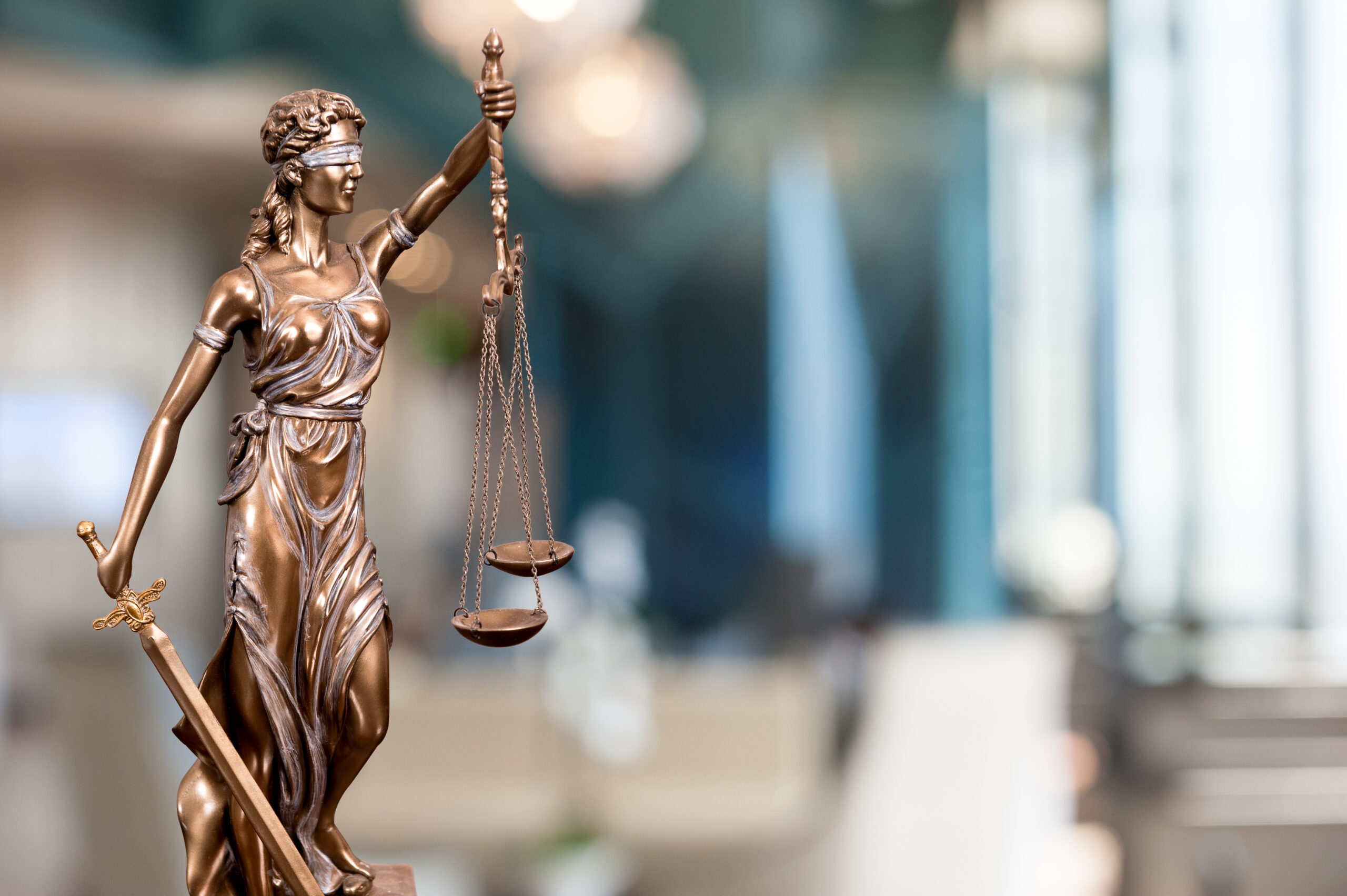 Bronze Lady Justice statue holding balanced scales and a sword, blindfolded, set against a blurred office background.