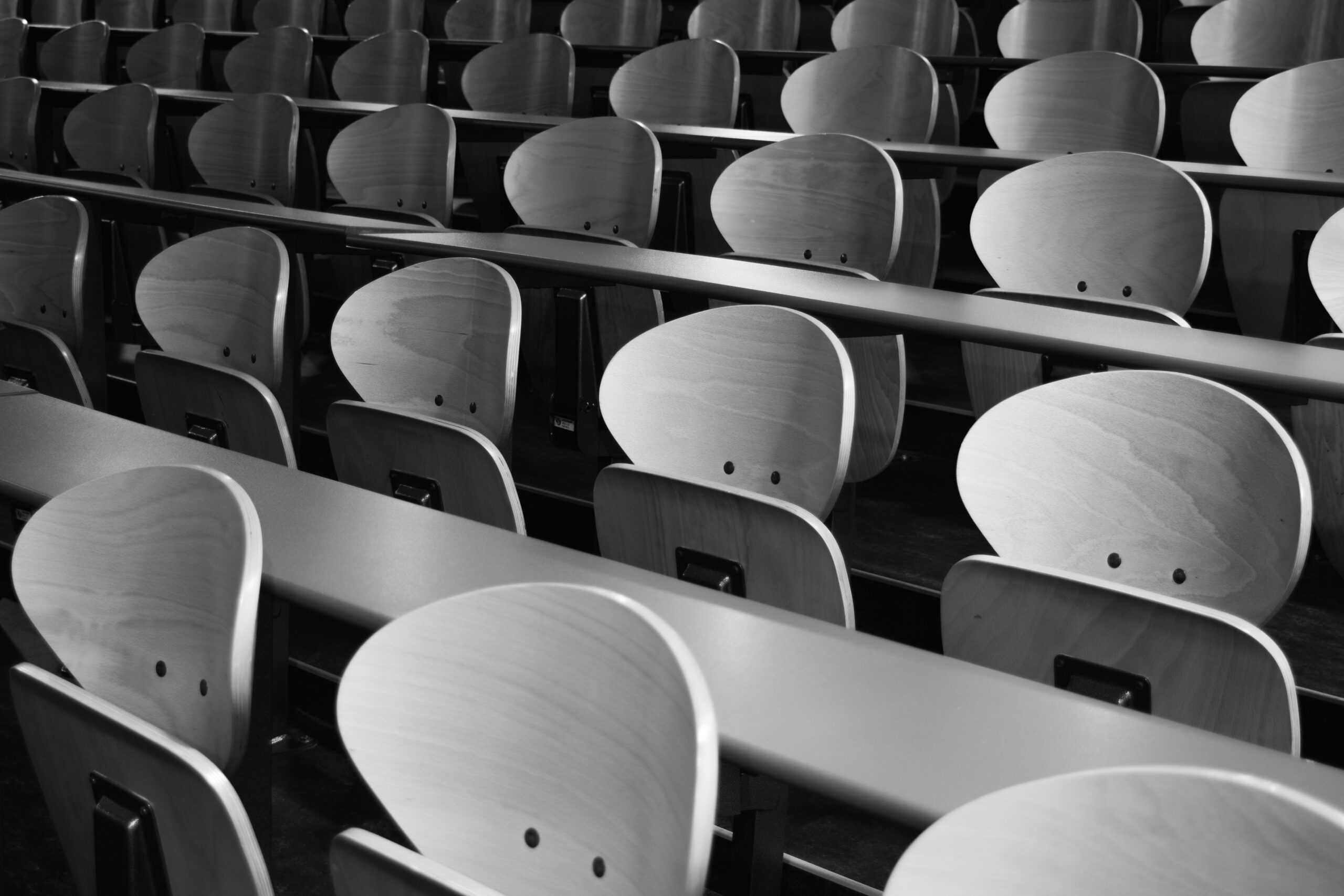 Rows of empty wooden chairs with curved backs in a lecture hall or auditorium, photographed in black and white