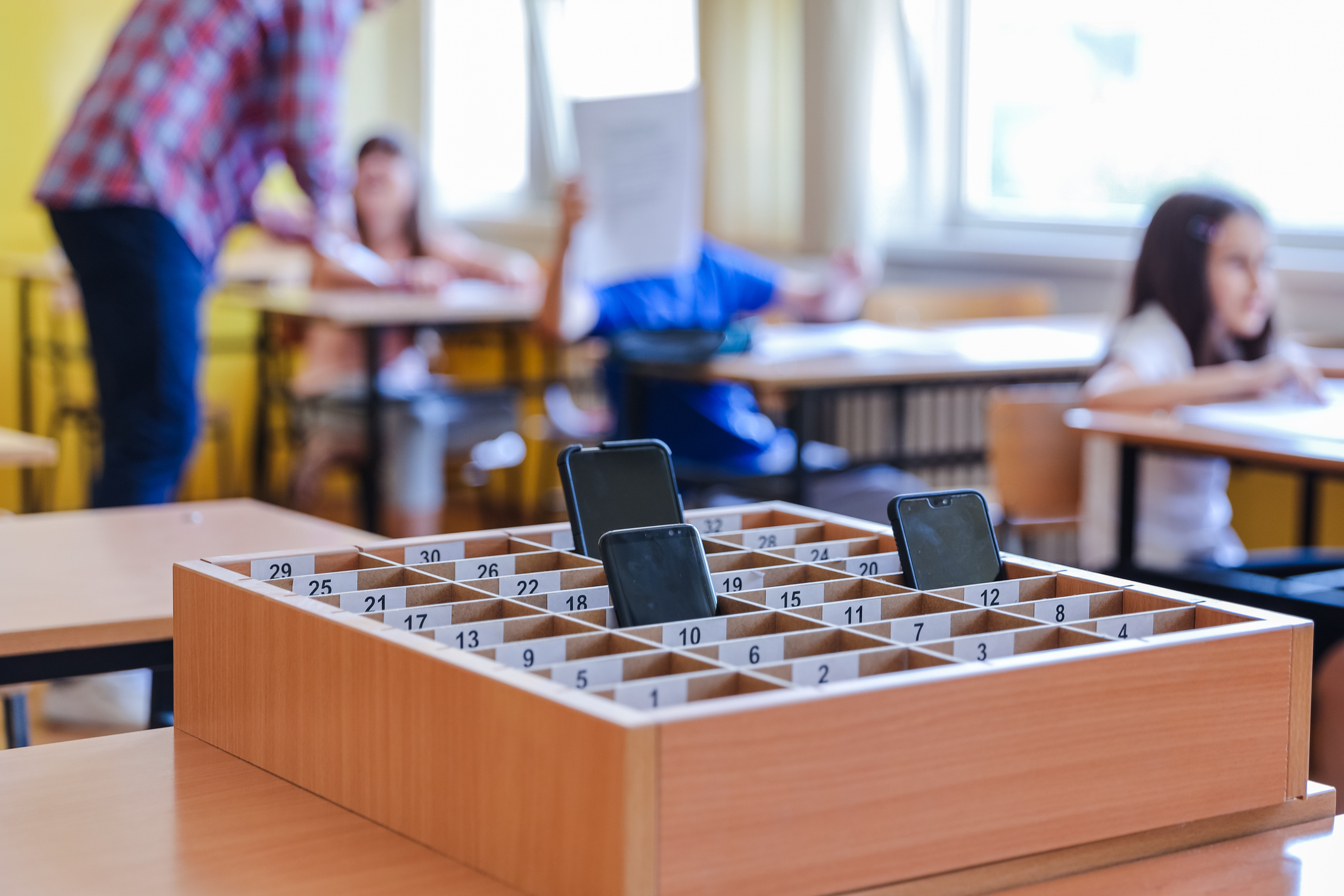 Numbered wooden phone storage box with two smartphones on a classroom desk, students and teacher visible in the blurred ba...