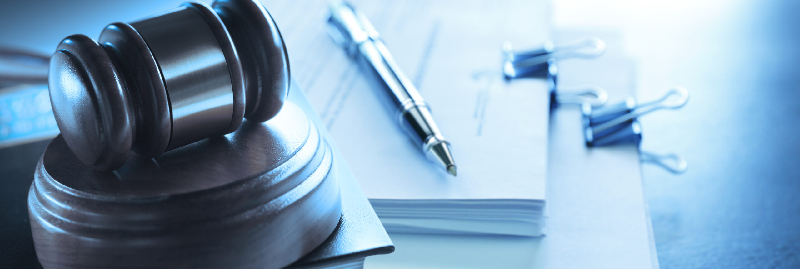 Wooden judge's gavel on sound block beside stacked legal documents, a pen, and binder clips on a desk with blue toning