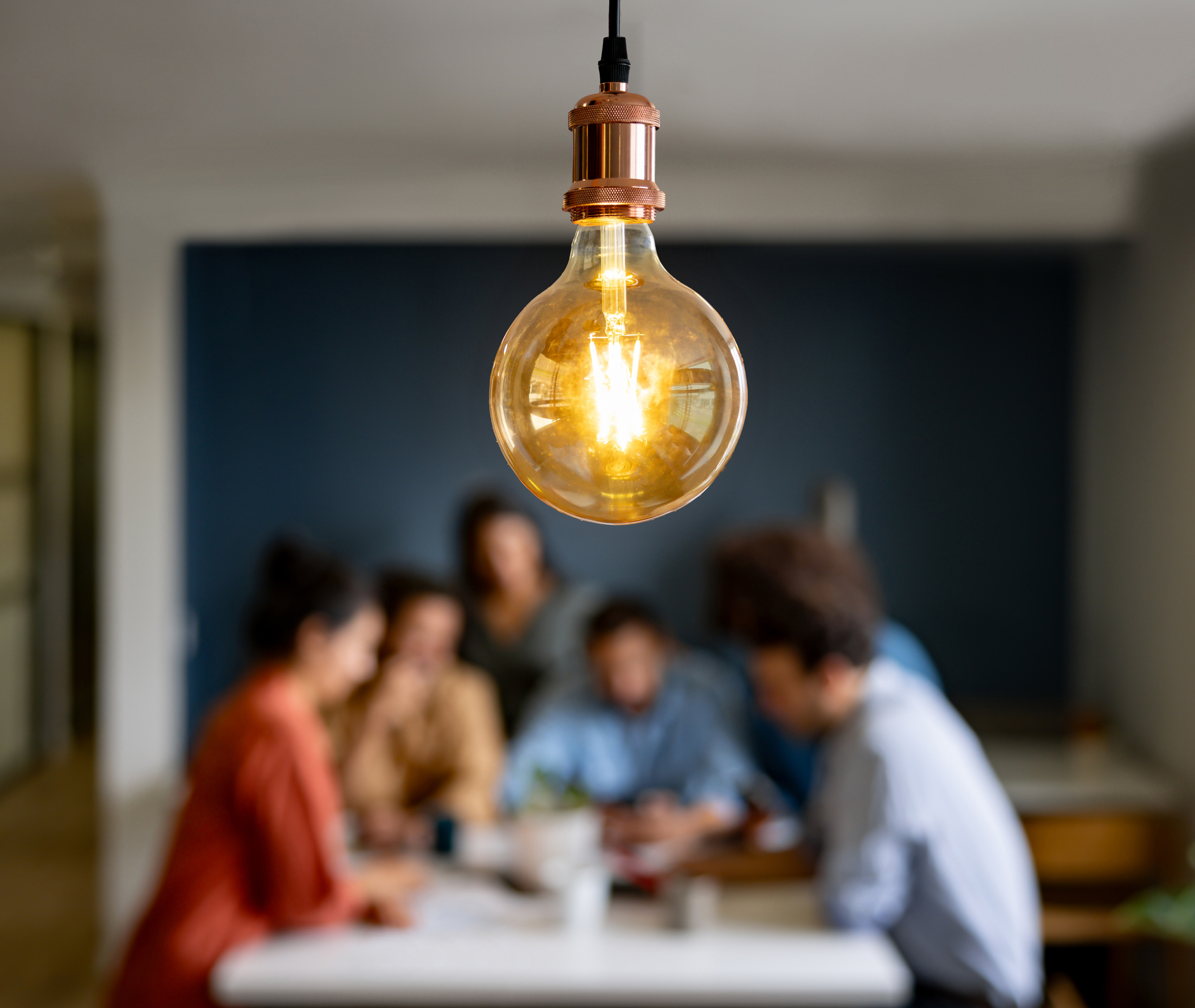 Glowing Edison bulb with copper fitting hanging above a blurred group of four people collaborating at a table
