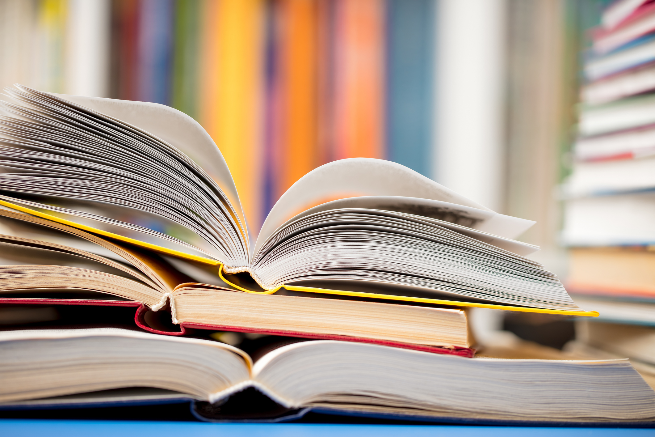 Open hardcover books stacked on each other with fanned pages, colorful books blurred in background on shelves