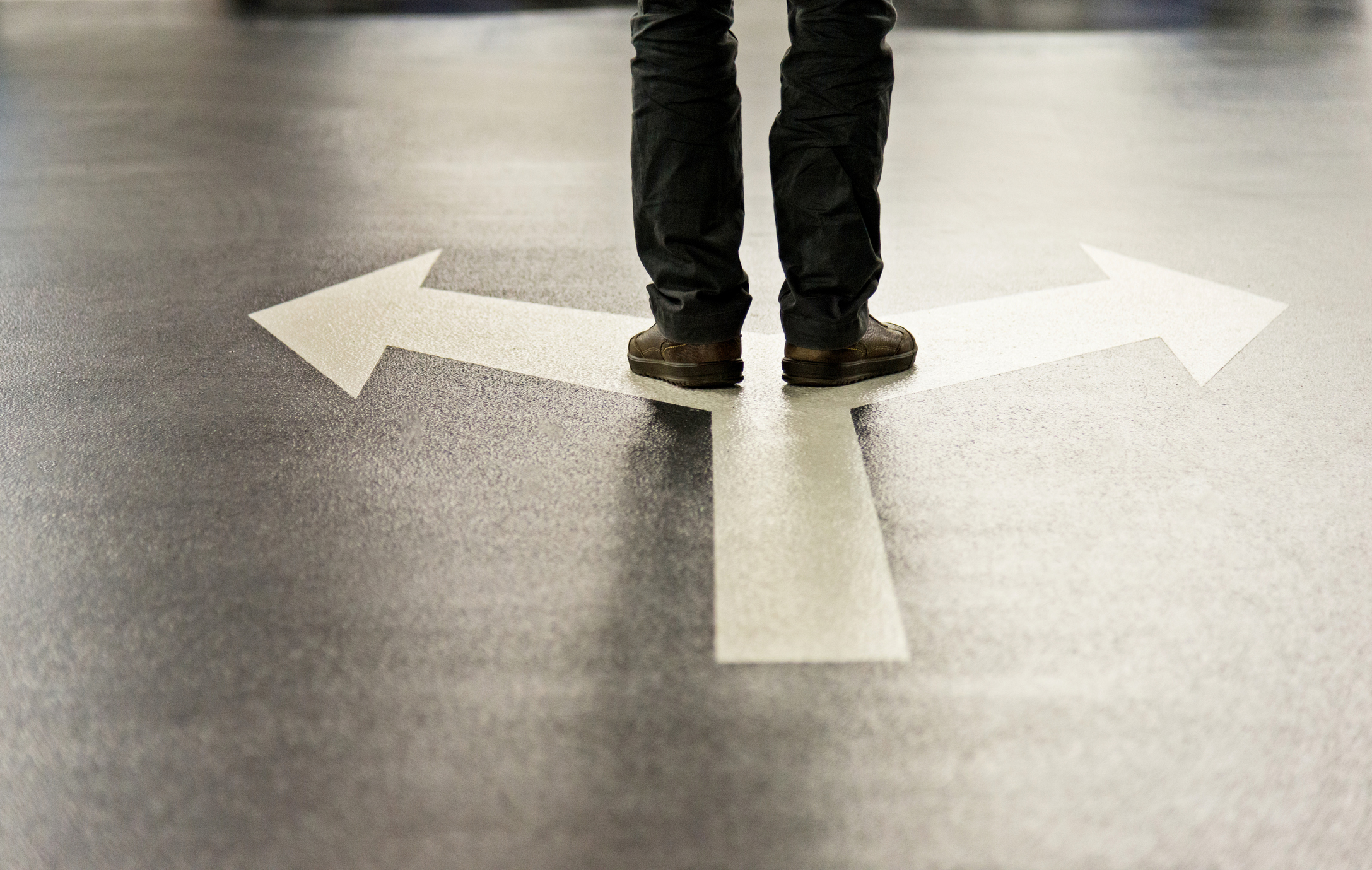 Person standing at a crossroads with three white arrows pointing left, right, and forward painted on a gray floor