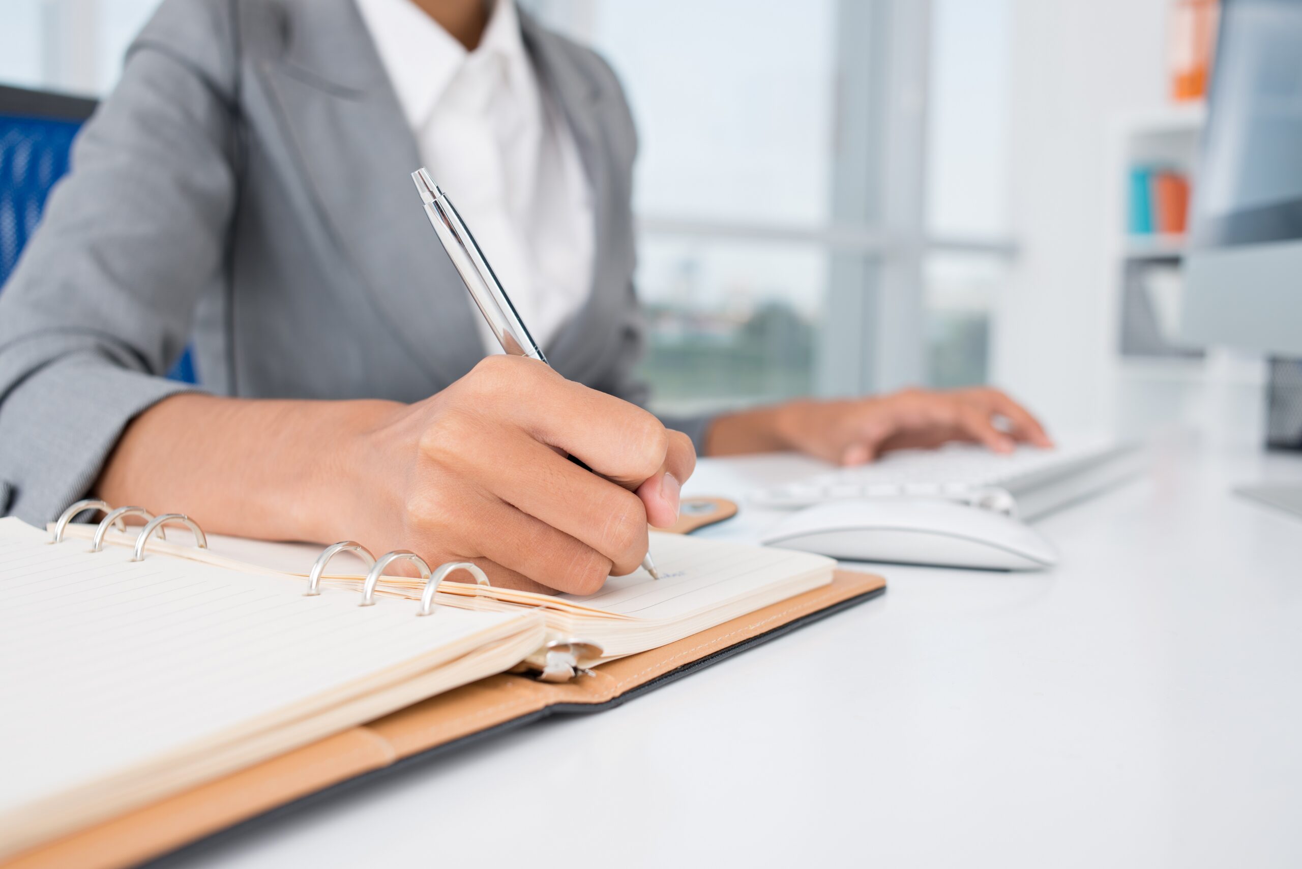 Professional woman in gray blazer writing in spiral notebook at desk while using a computer keyboard and mouse